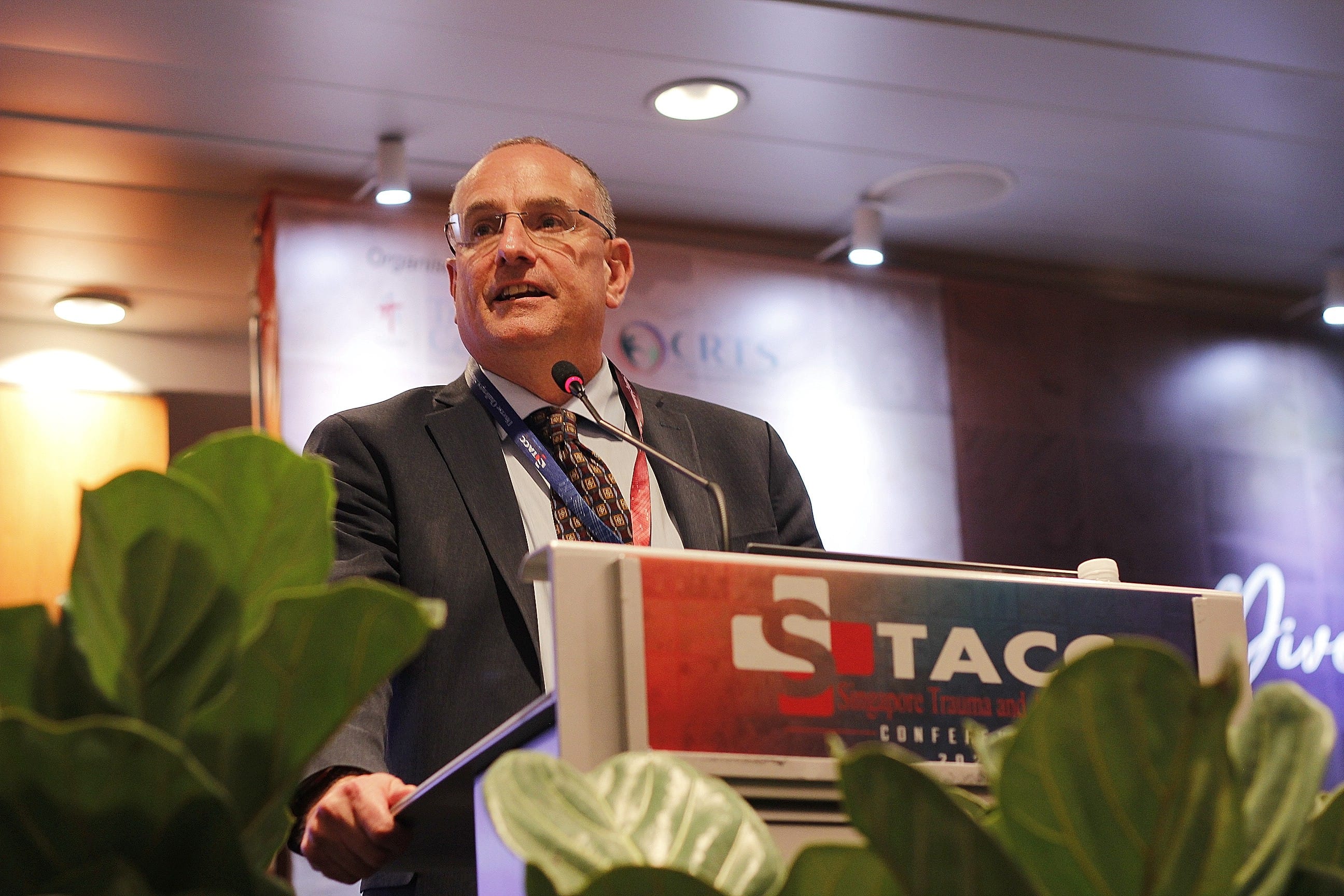 Man in suit and glasses speaking at a podium labeled Singapore Trauma and Acute Care Conference.