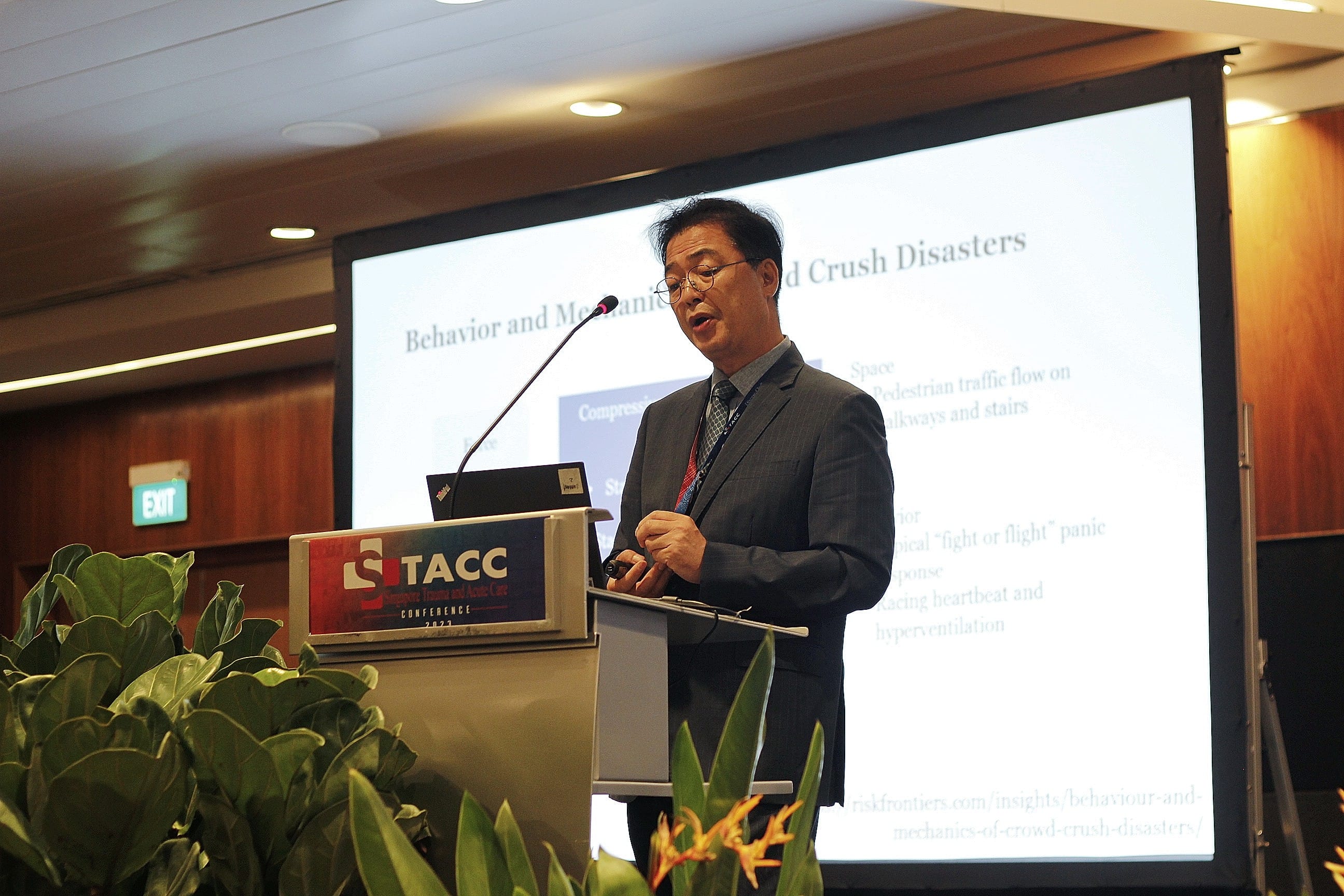 A man in a gray suit speaks at a STACC Conference podium with a slide about Crush Disasters on screen behind him.