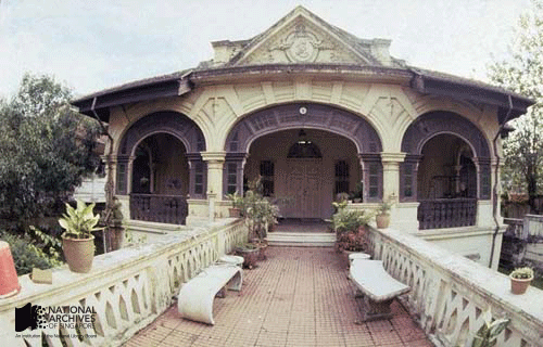 Colonial-style building with arched entrance, decorative facade, white balustrade, potted plants, and benches on a tiled terrace.