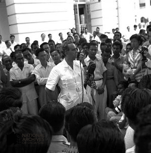 A man in a white shirt speaks into a microphone before a large crowd outdoors in a black and white photo.