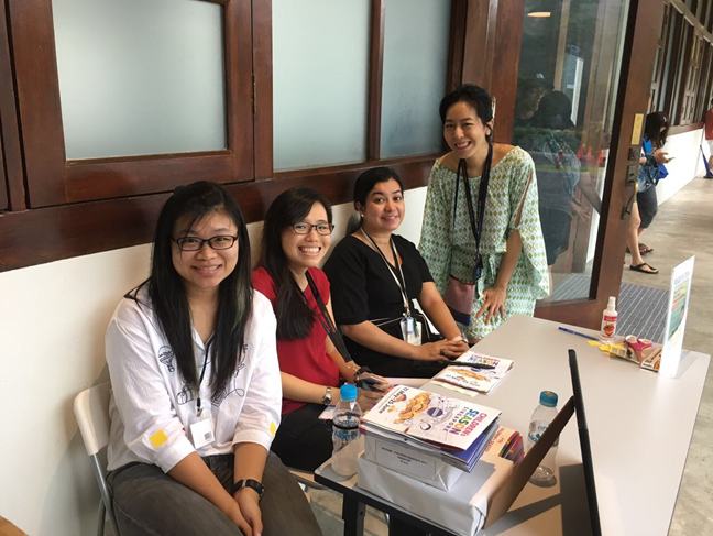 Four women smile at a registration or welcome table with books, badges, and water bottles indoors.