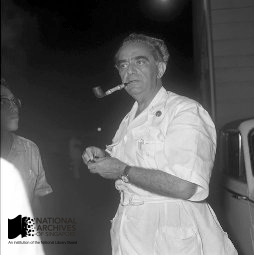 Black and white photo of a middle-aged man in a white shirt smoking a pipe outdoors.
