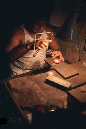 A man in a white tank top works by dim light at a wooden table, holding a small tool over craft materials.