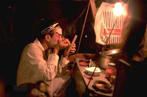 A performer applies white face makeup by candlelight at a dimly lit backstage dressing table.