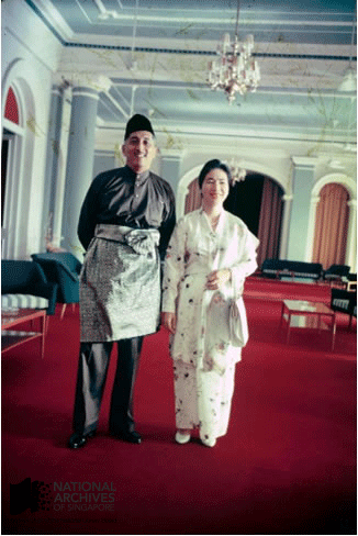 A man in traditional Malay attire and a woman in a floral kimono stand together in an ornate room with red carpet.