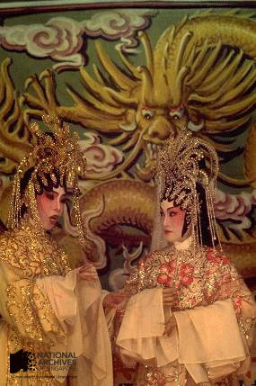 Two Cantonese opera performers in ornate gold costumes and headdresses stand before a painted dragon backdrop.