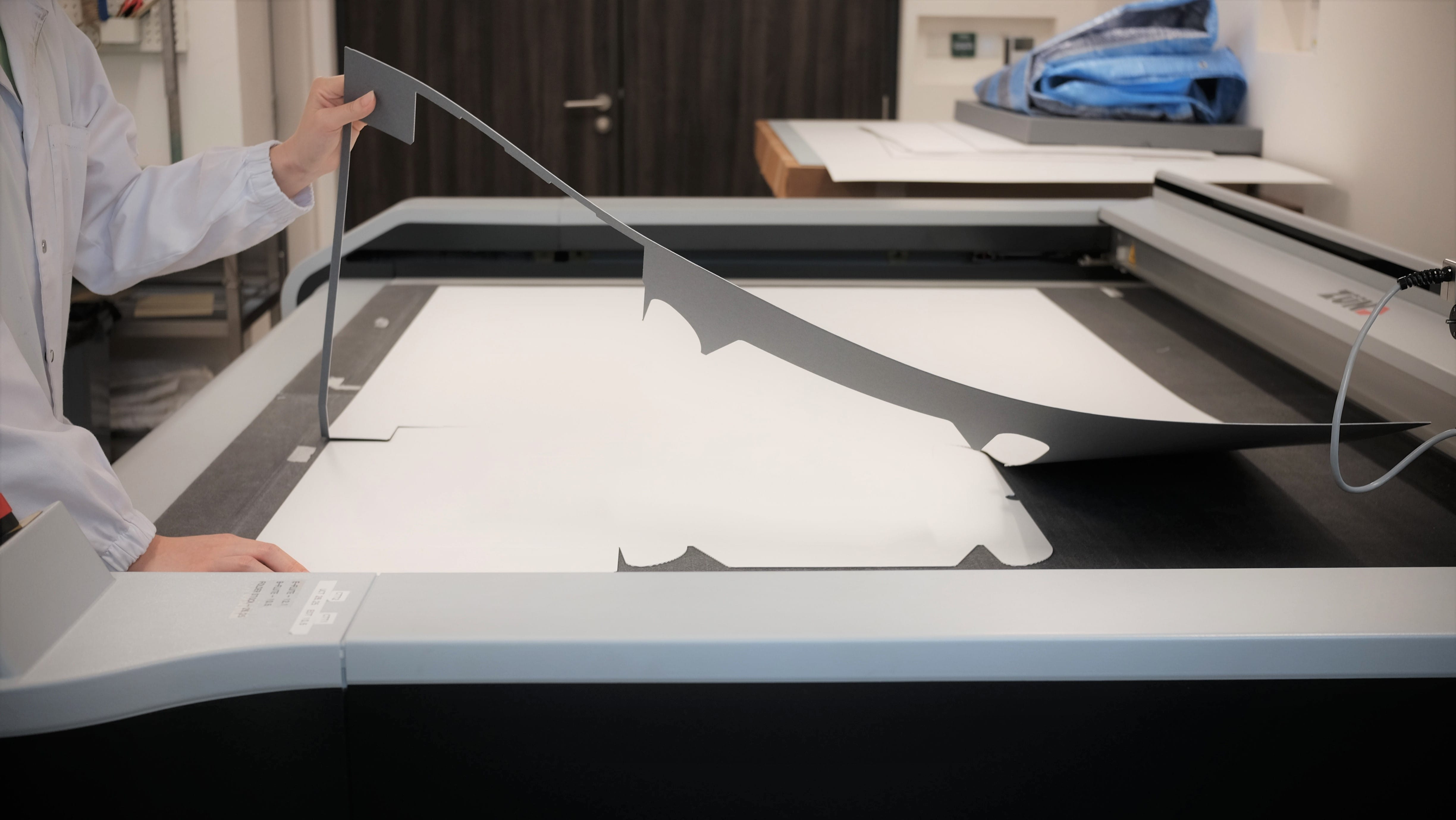Person in a white coat peeling a cut dark sheet from a large flatbed cutting machine.
