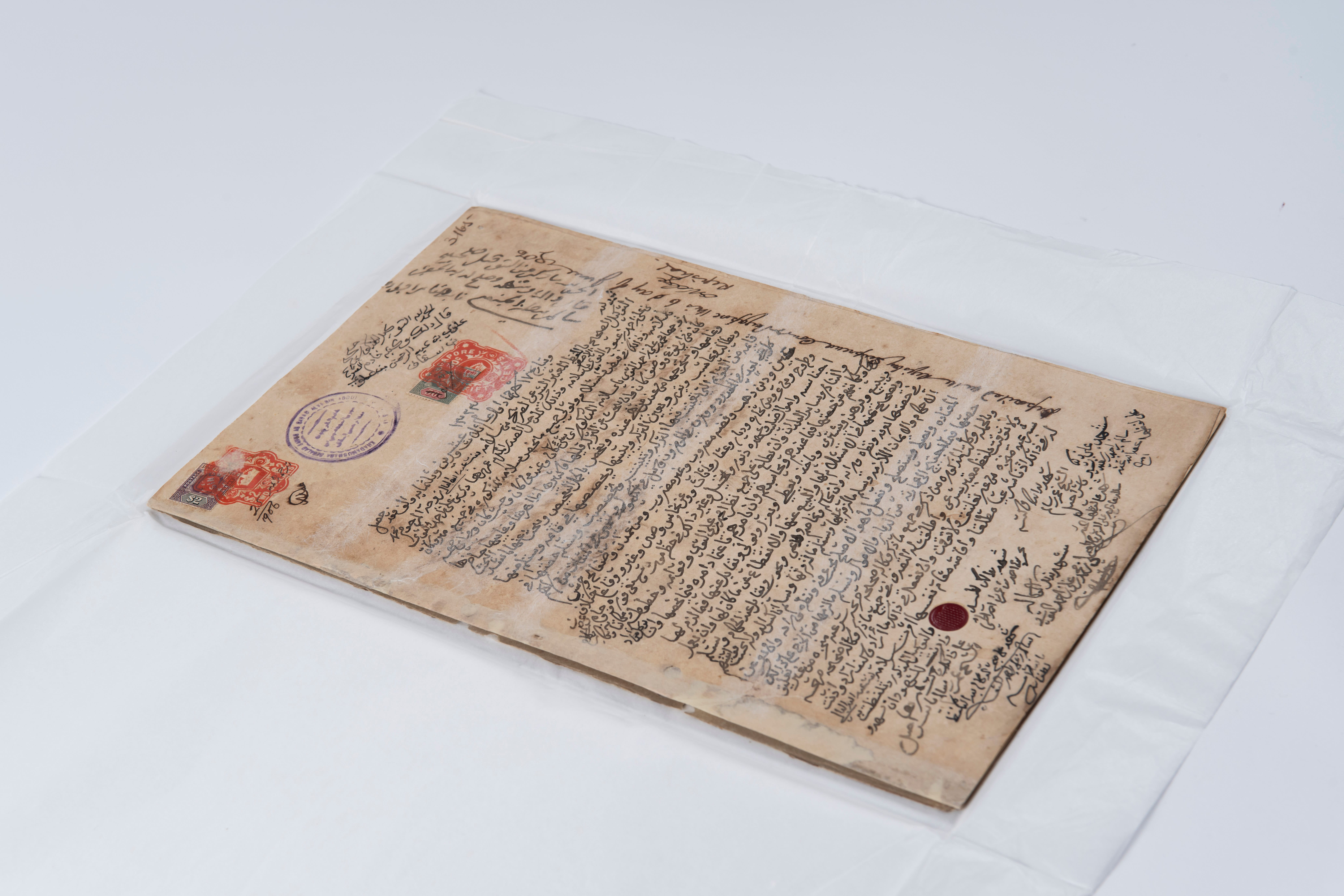 Aged handwritten document in a clear sleeve, with Arabic script, stamps, and a red wax seal.