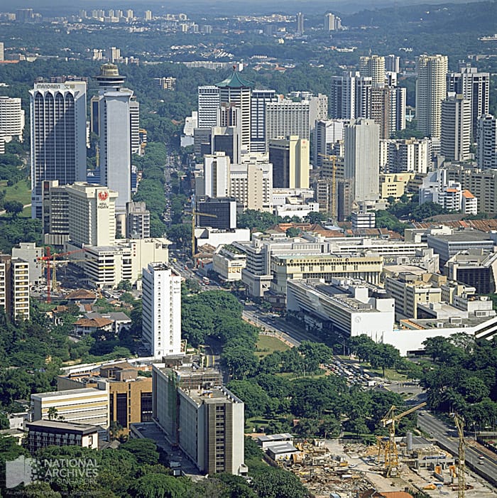 Aerial view of a dense urban skyline with tall buildings, tree-lined streets, and construction cranes visible.