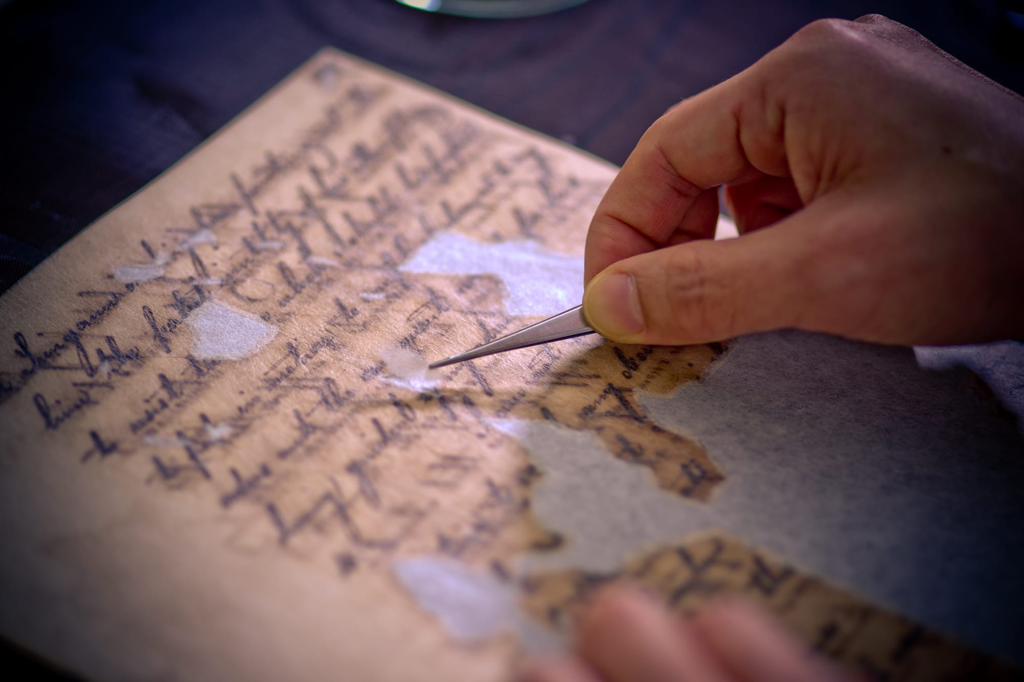 A hand uses tweezers to carefully repair a fragile aged document covered in handwritten cursive text.