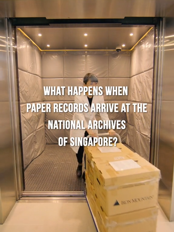 A woman pushes a cart of stacked archive boxes out of a padded elevator with text about the National Archives of Singapore.
