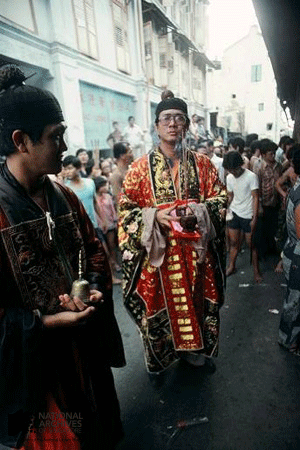 Two men in ornate traditional Chinese robes walk in a crowded street during a cultural procession.