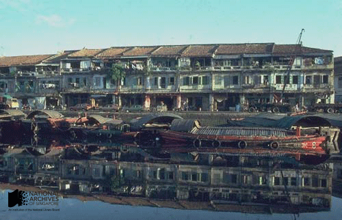 Old shophouses and wooden boats reflected in calm river water along a Singapore waterfront.