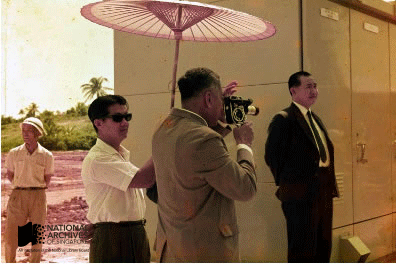 Three men standing outdoors, one filming with a camera, under a pink parasol, near a building.