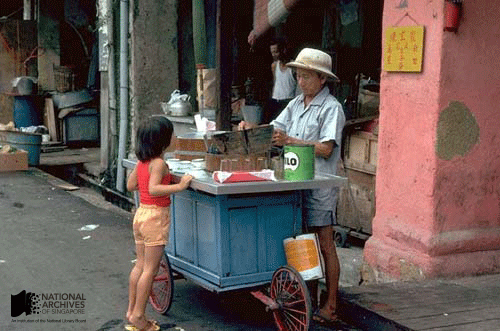 An elderly street vendor with a hat serves a young child at a blue wheeled cart on an old urban street.