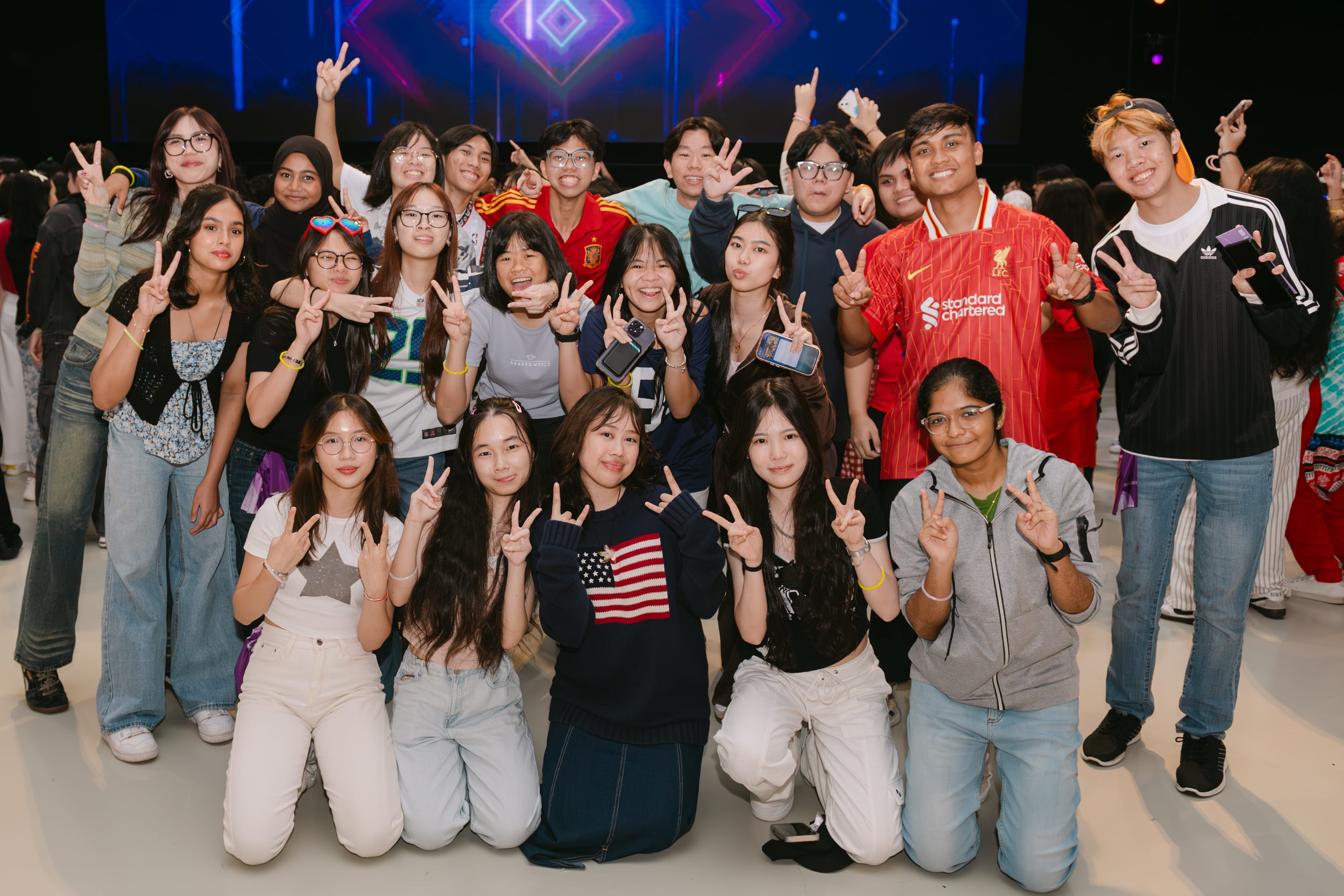 A large group of young people posing with peace signs in front of a lit stage backdrop.