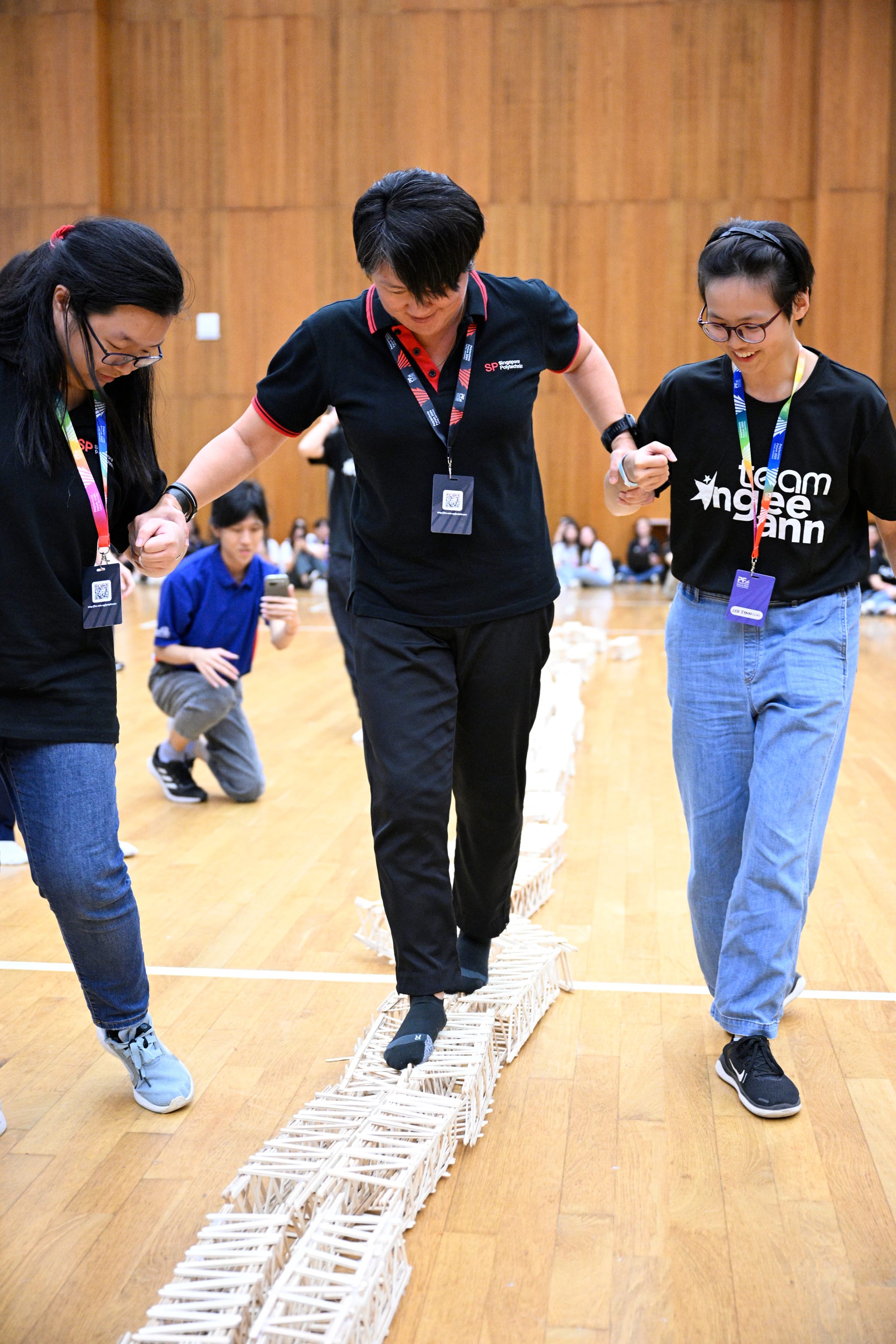 Three people holding hands walk carefully along a narrow popsicle stick bridge on a gymnasium floor.