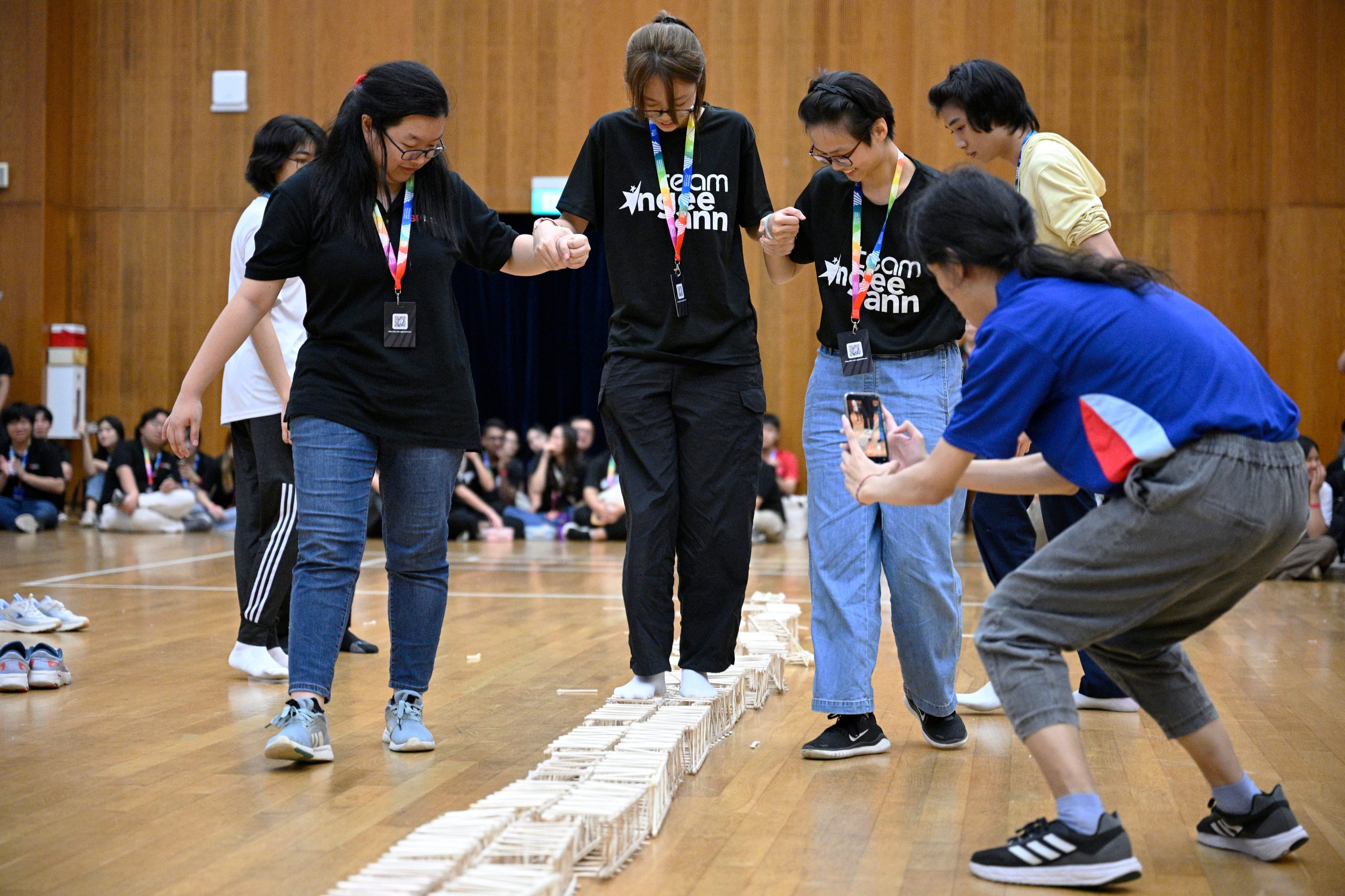 Students in a gym carefully walk along a long wooden popsicle stick structure on a polished floor.