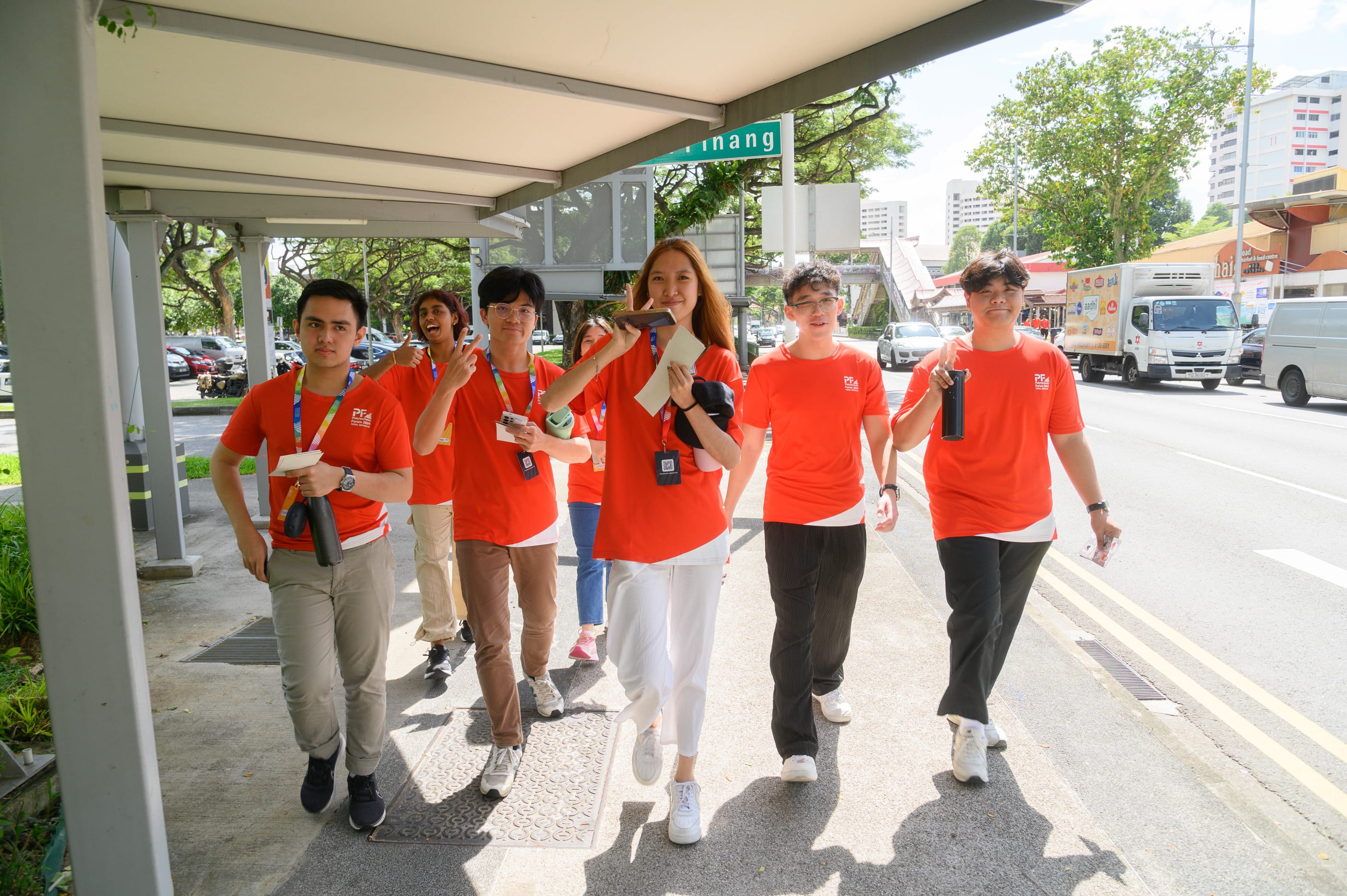 A group of young people in matching red T-shirts and lanyards walking along a covered sidewalk near a busy road.