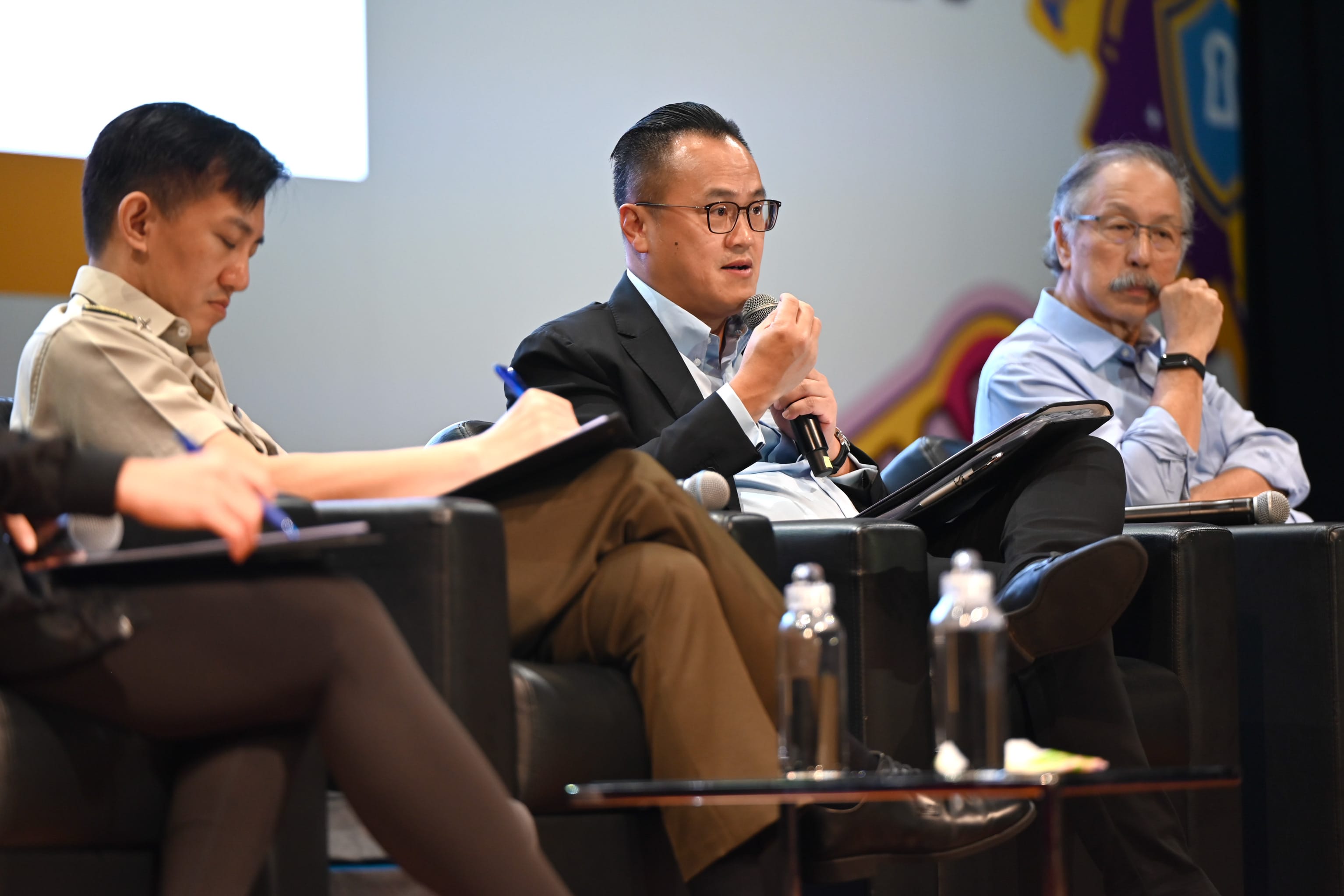 Three people sit in black chairs on a panel; the center man speaks into a microphone while others listen.