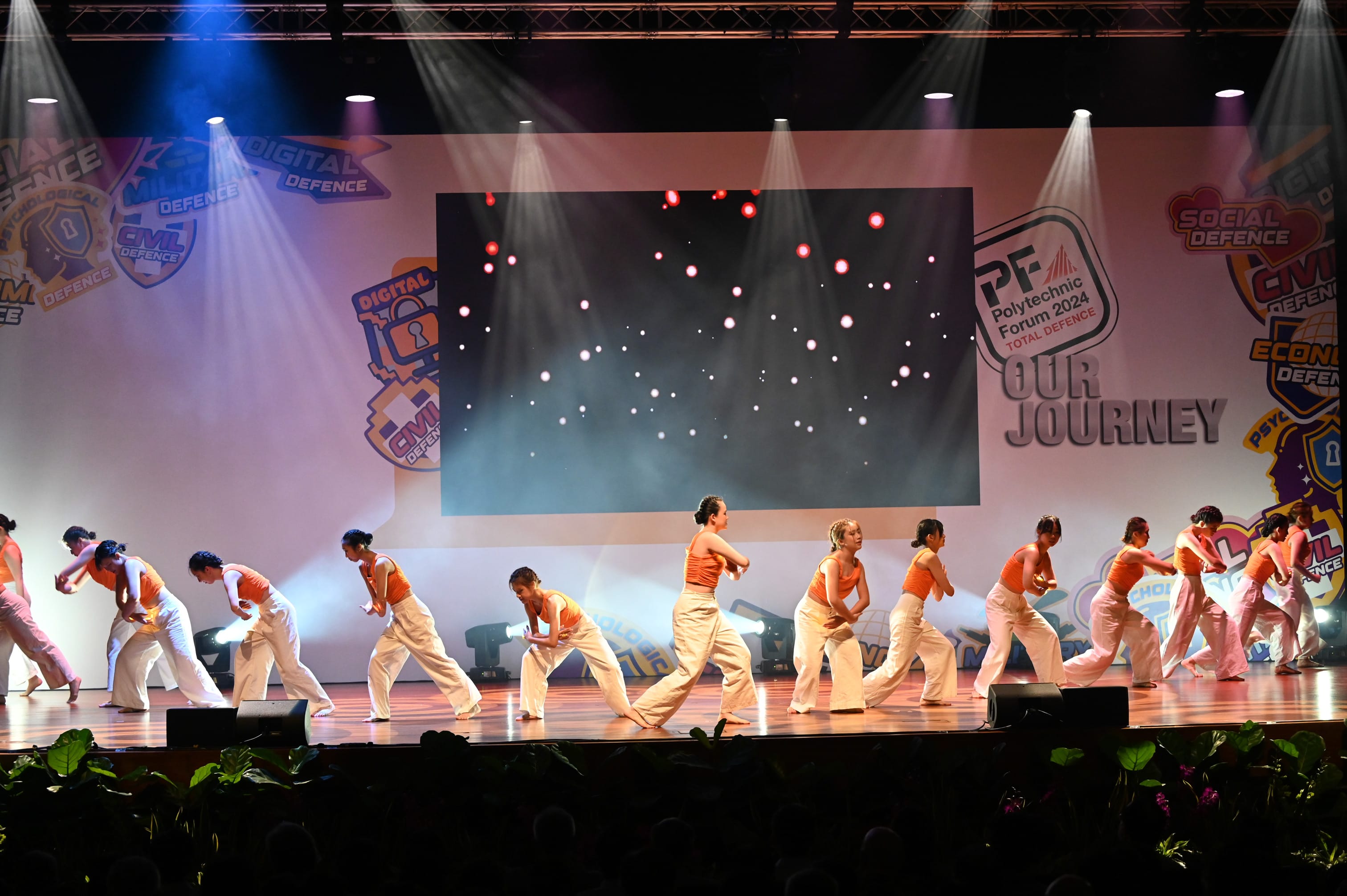 Dancers in orange tops and white pants perform on a lit stage at Polytechnic Forum 2024 Total Defence event.