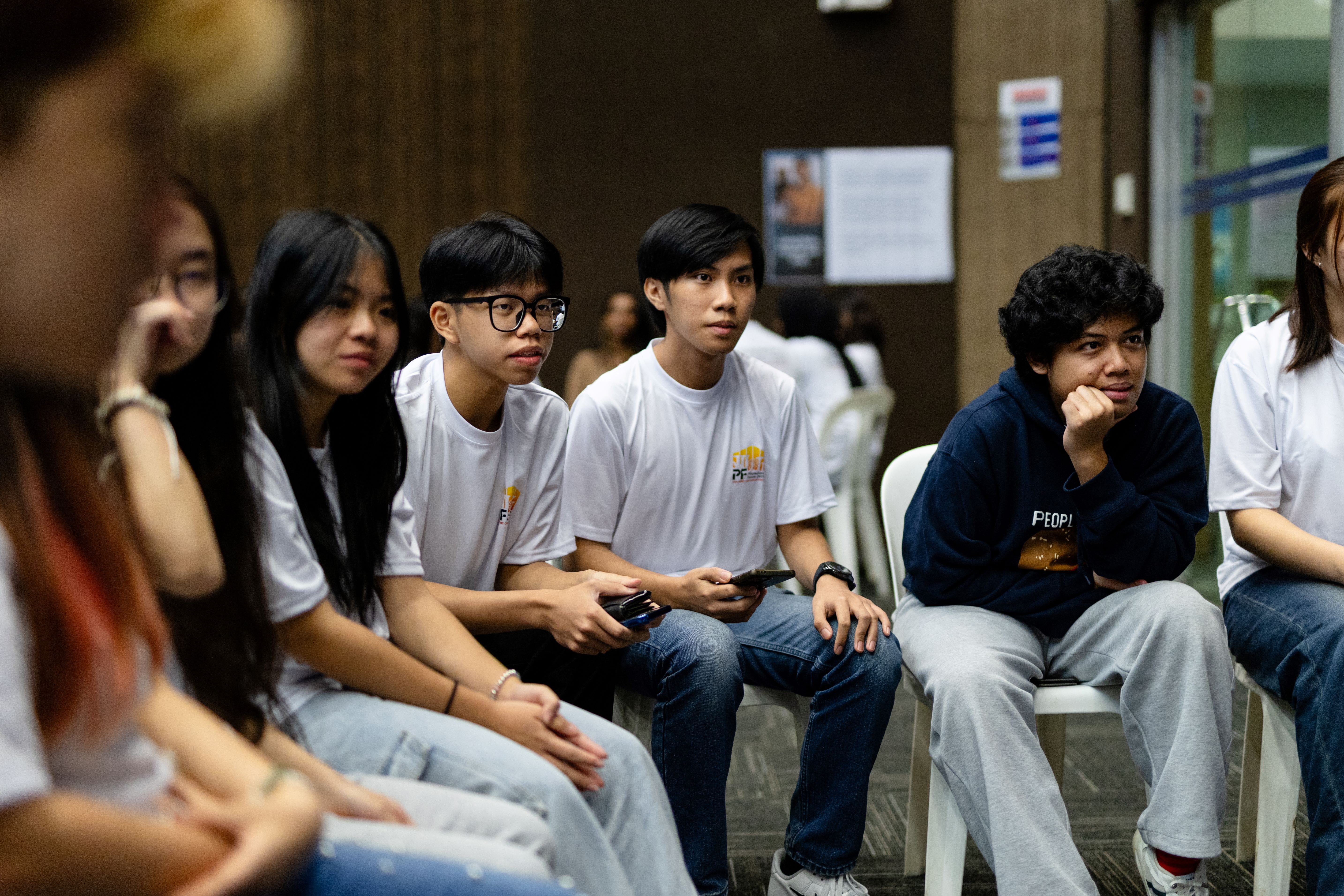 A group of young people sit on white chairs attentively listening during an indoor gathering or event.