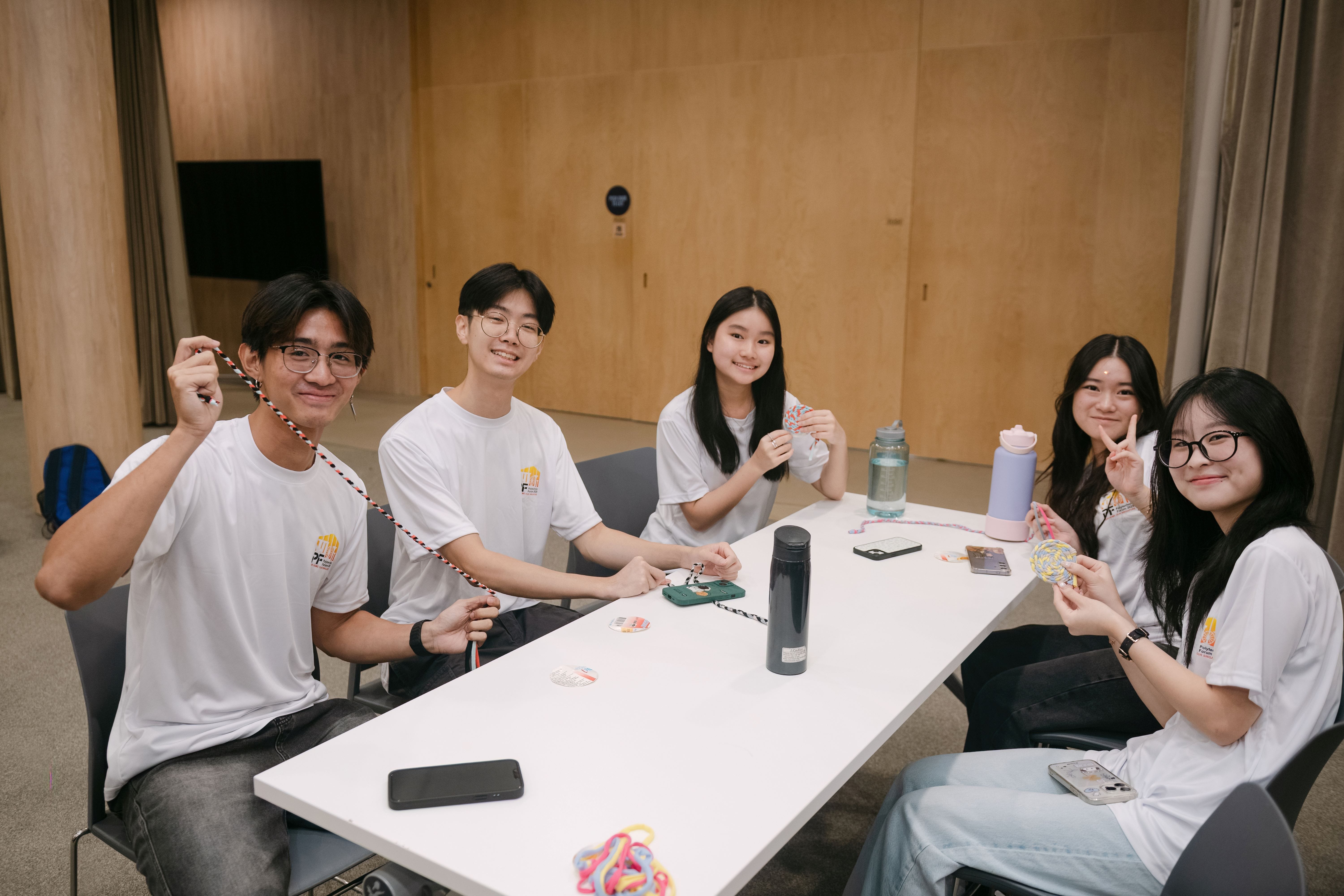 Five young people in white shirts sit at a table, smiling and holding handmade crafts like braided cords and colorful ornaments.