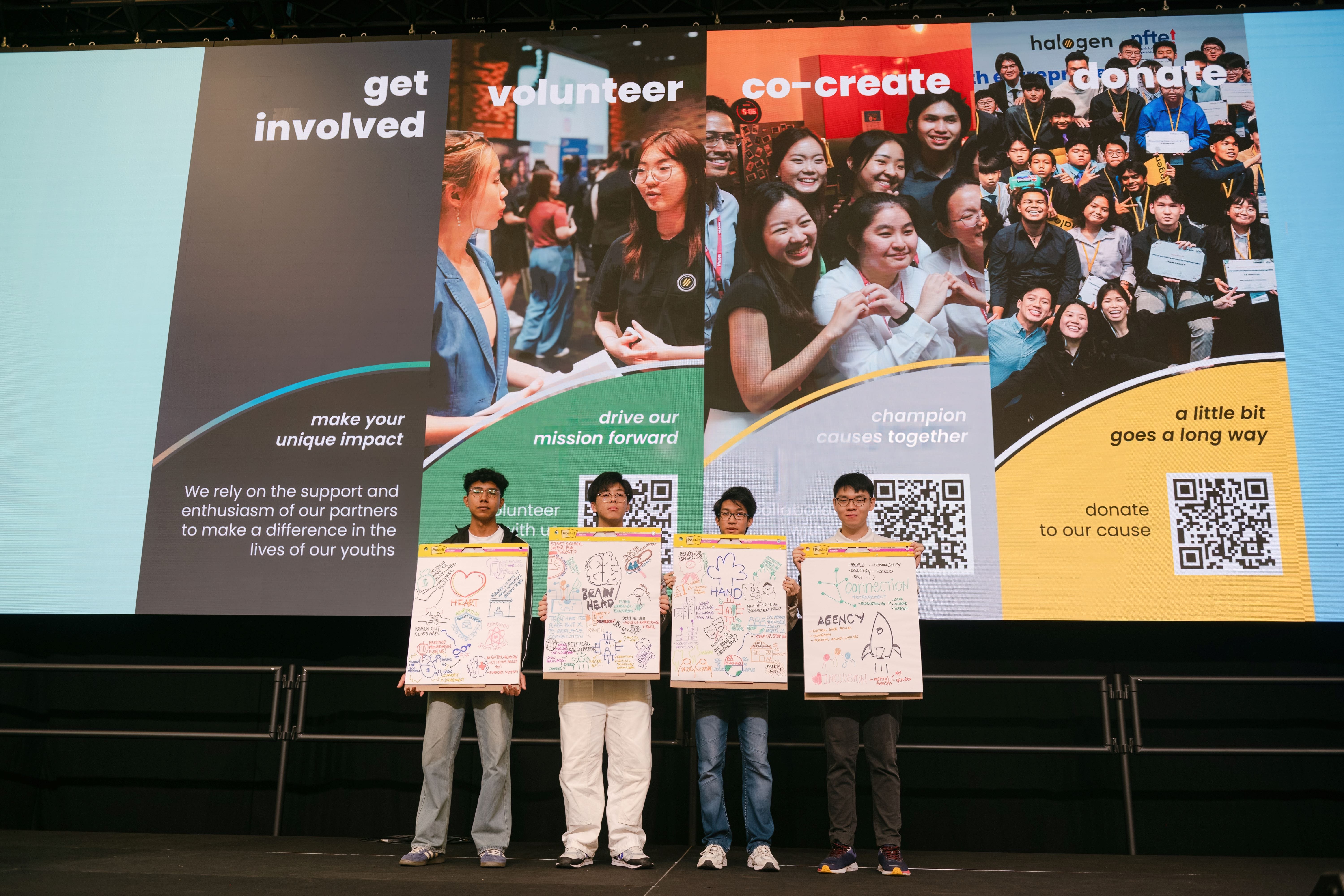 Four young people stand on a stage holding hand-drawn poster boards in front of a large screen with volunteer and donation messages.