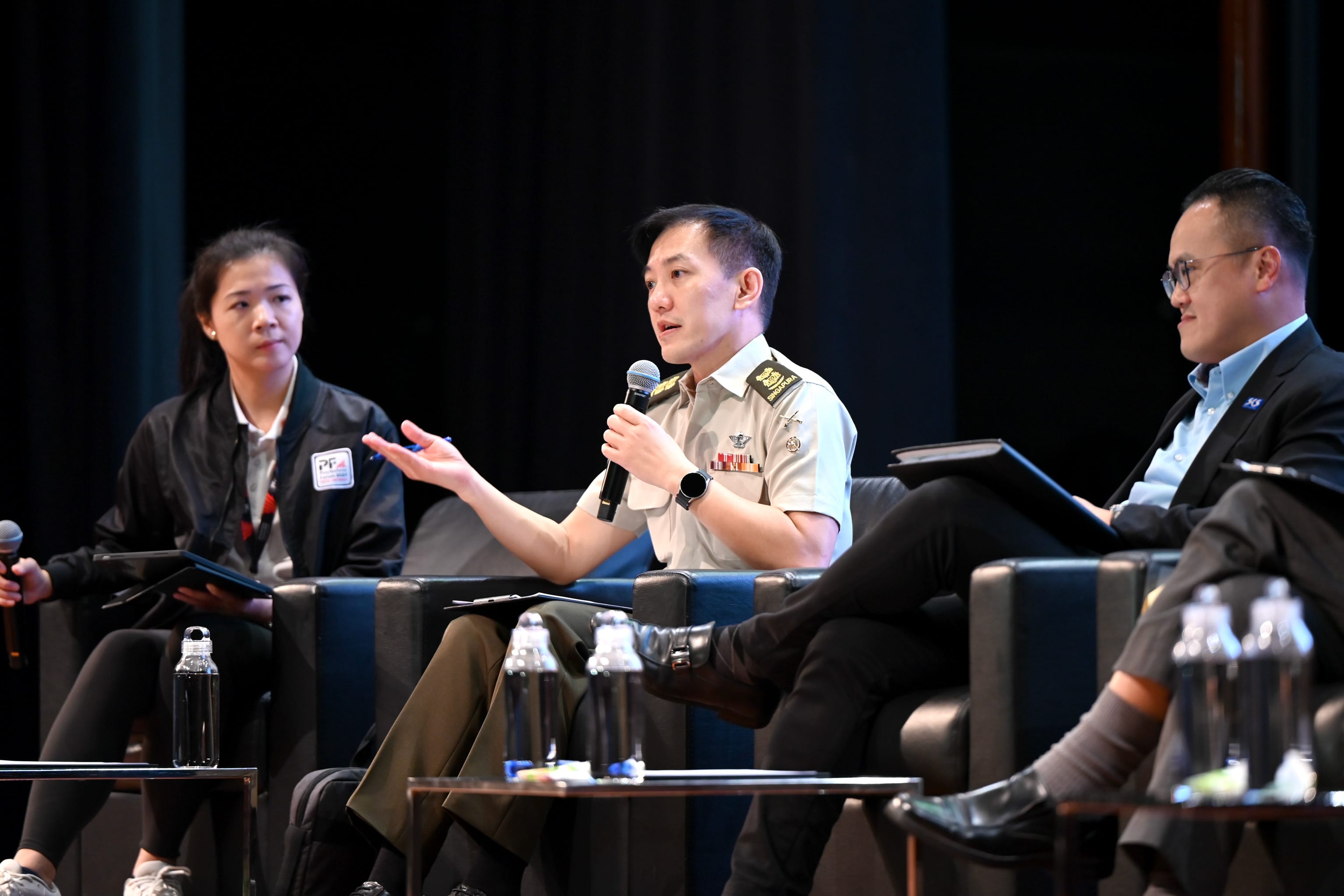 Three panelists seated on stage; a uniformed military officer speaks into a microphone while gesturing with one hand.