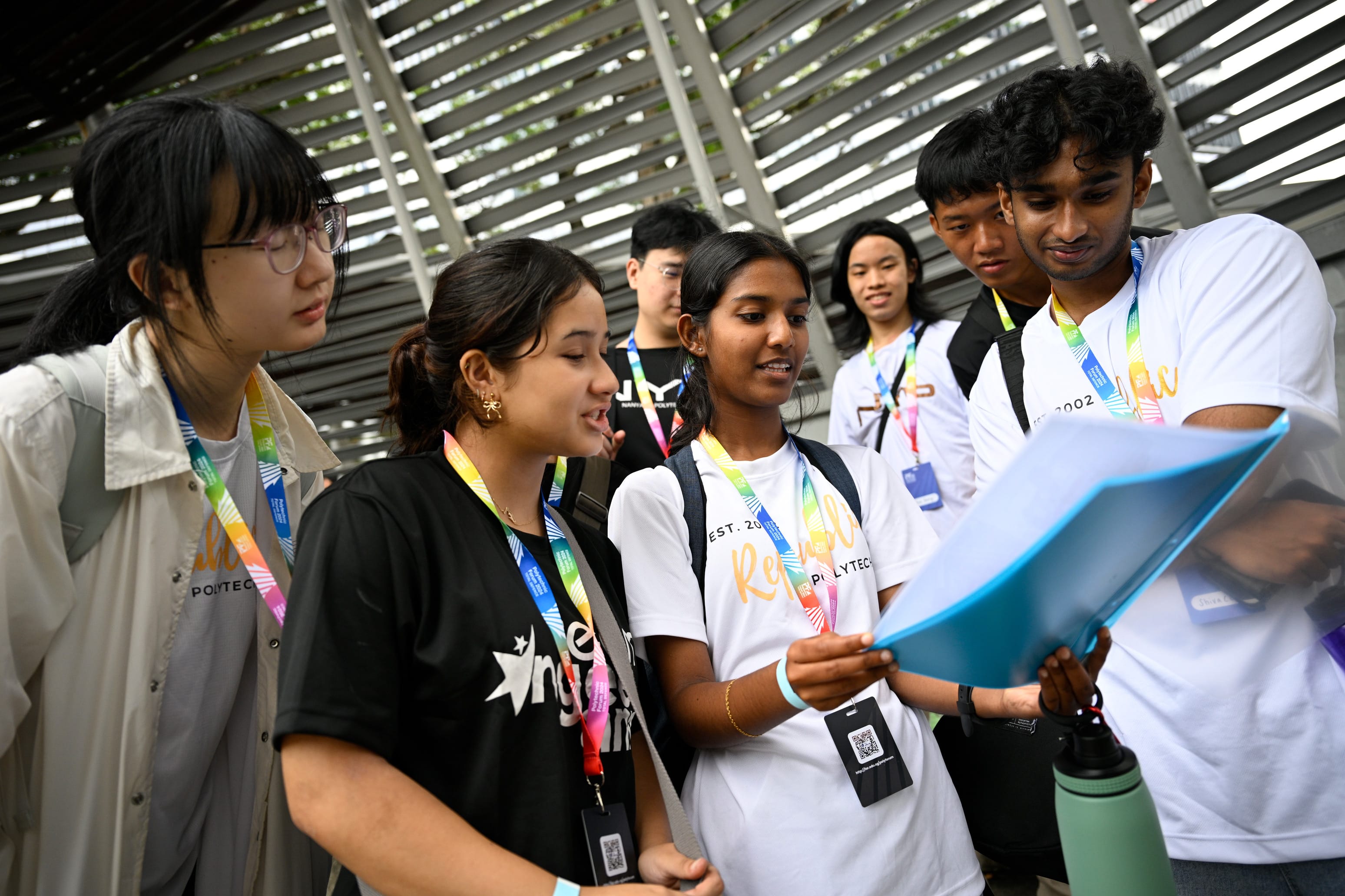 Six young students wearing colorful lanyards gather closely, looking at a blue tablet held by one student.
