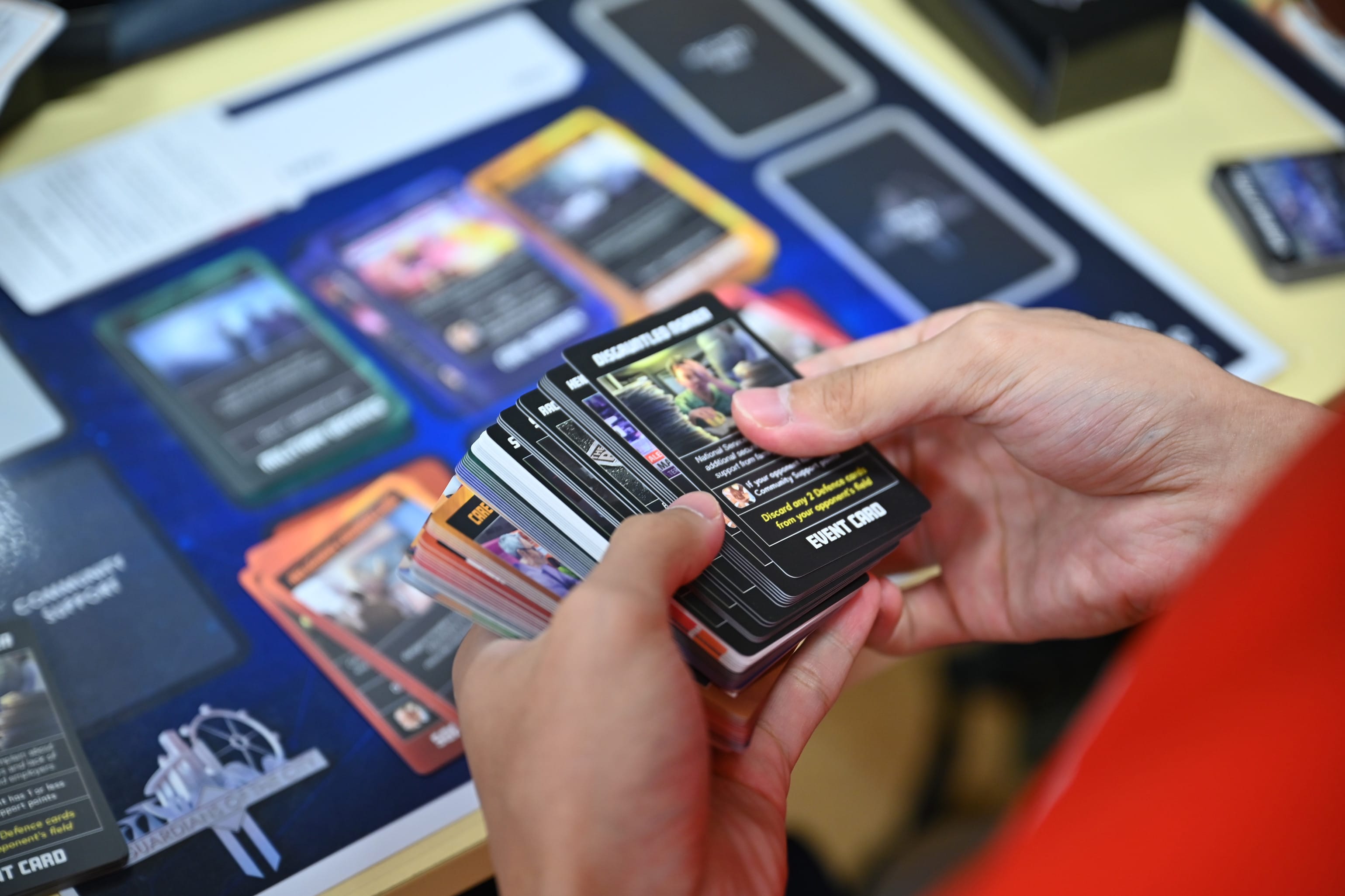 Hands holding a stack of dark trading cards labeled "Event Card" over a blue board game play mat with cards laid out.