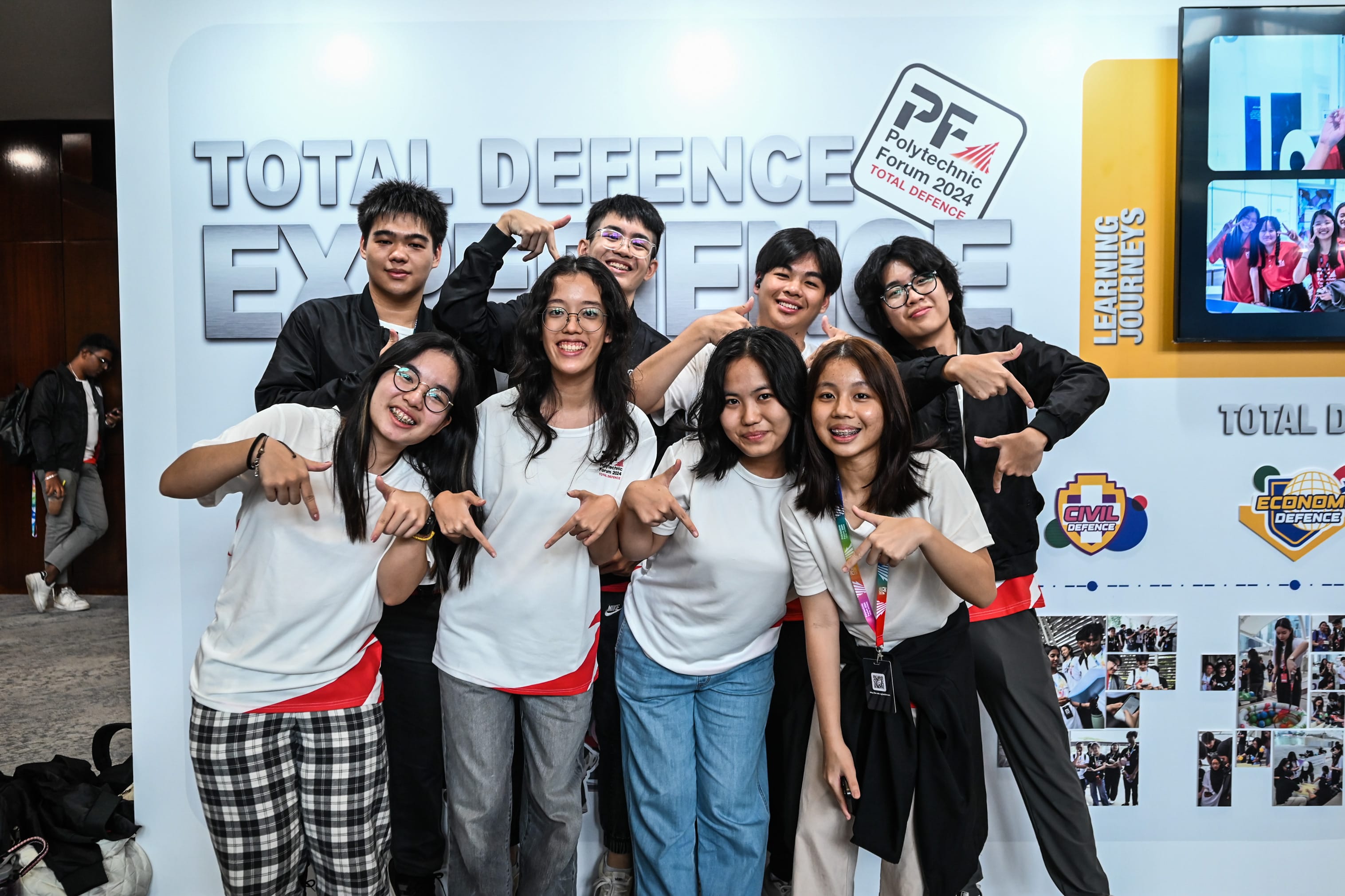 Seven smiling students pose in front of a Total Defence Experience banner at Polytechnic Forum 2024.