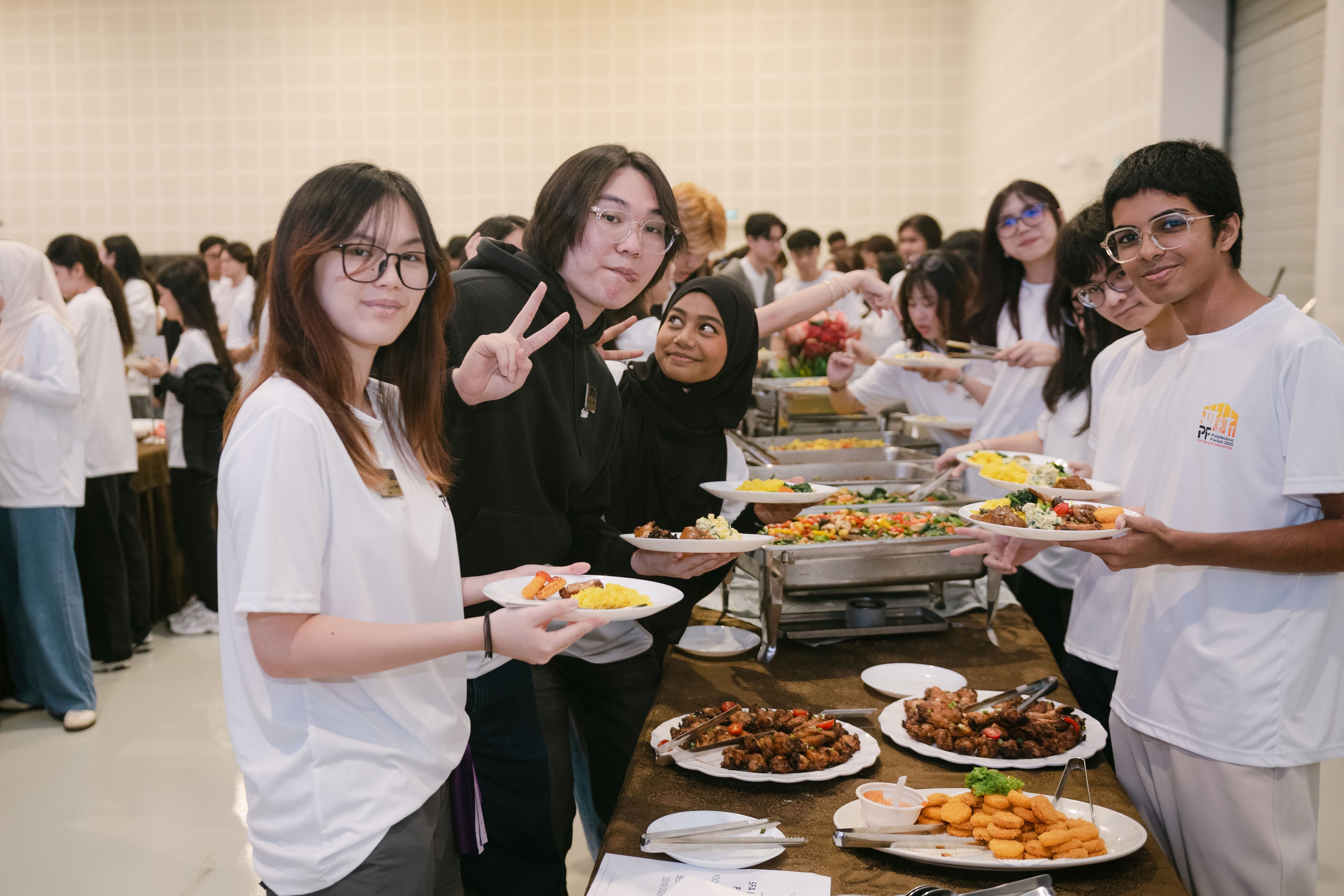 Students smiling and holding plates of food at a buffet line during a large indoor event.