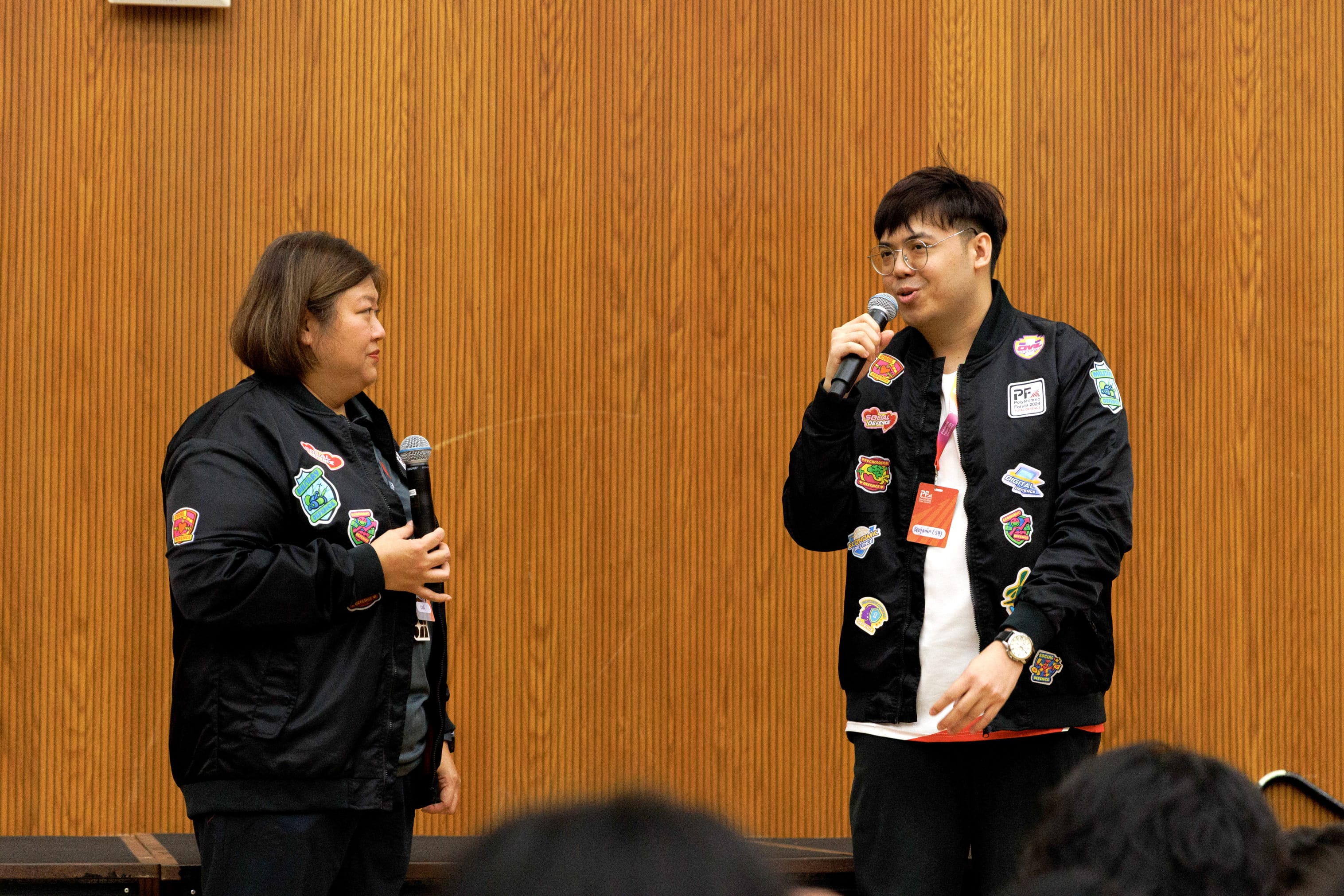 Two people in black jackets with colorful patches hold microphones and speak on stage before an audience.