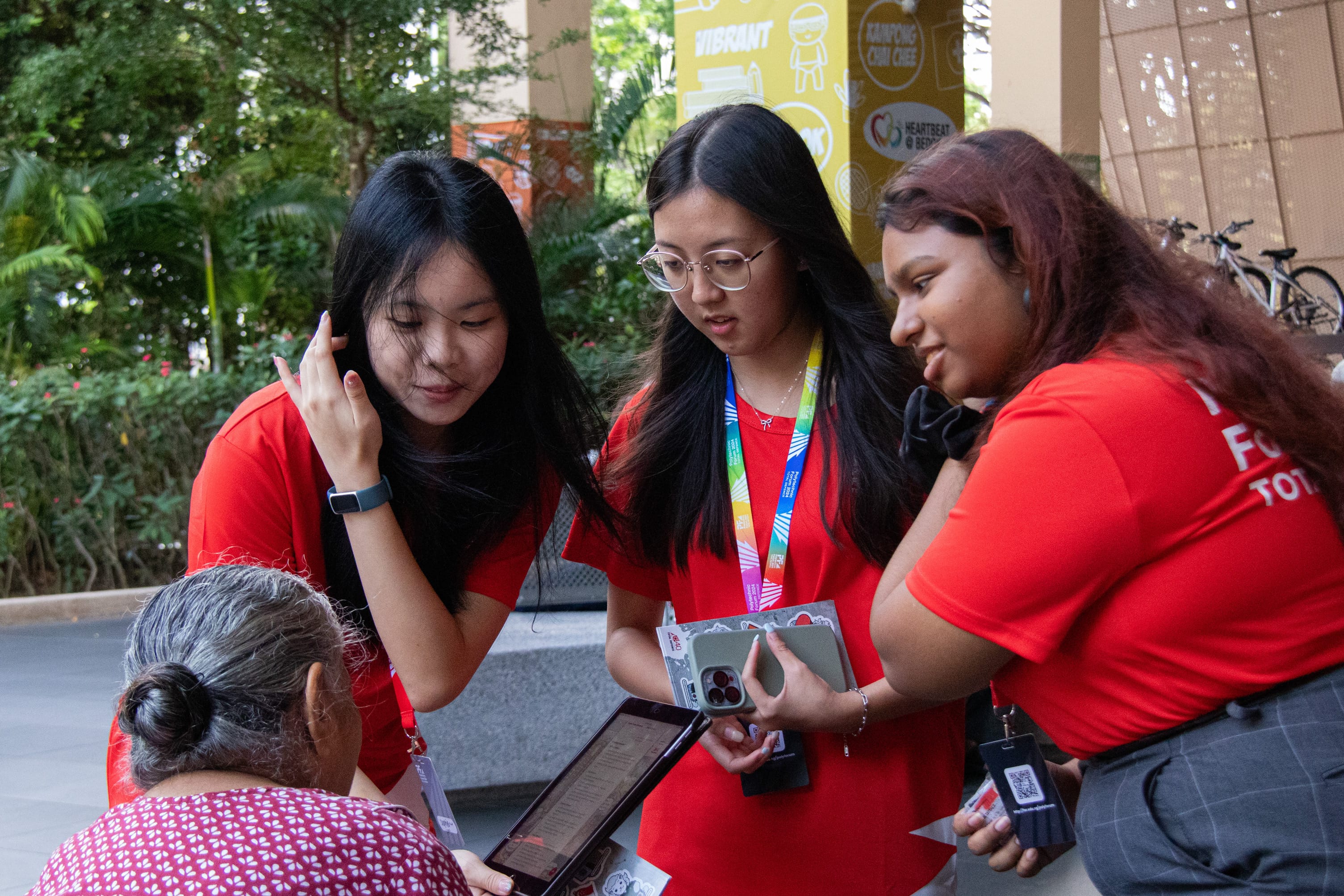 Three young women in red shirts show a tablet to an elderly woman outdoors near a yellow banner.