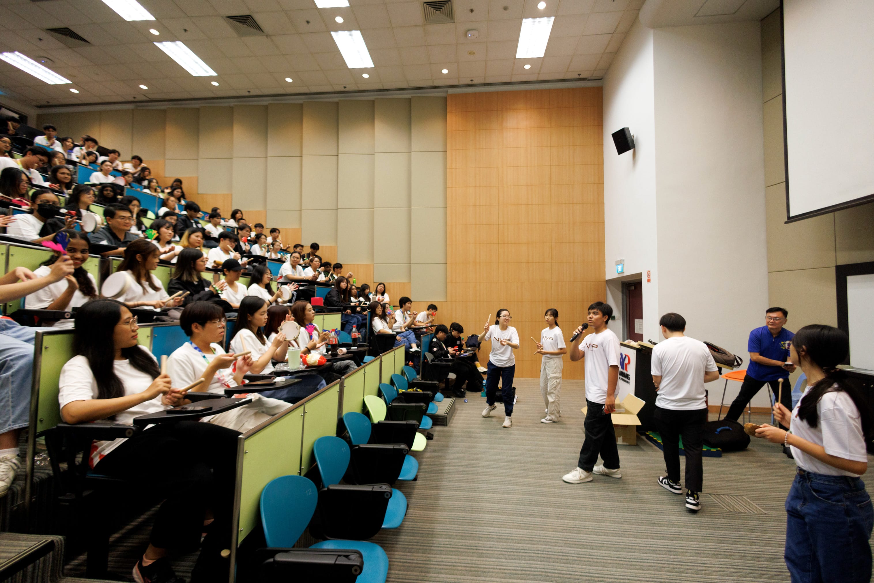 Students in white shirts fill a large lecture hall while a few stand at the front holding microphones and drumsticks.