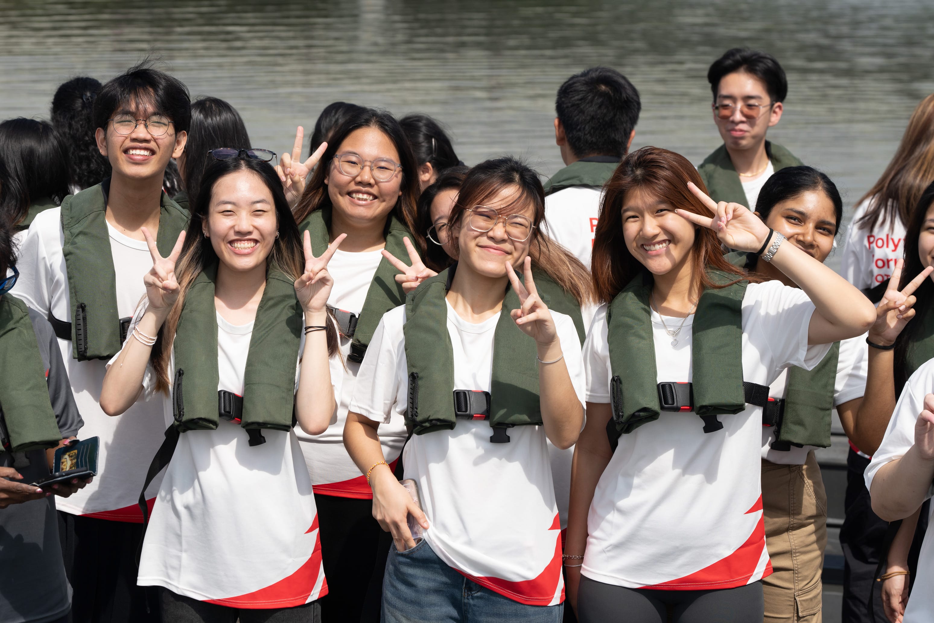 A group of smiling young people wearing green life jackets and white shirts pose with peace signs near a body of water.