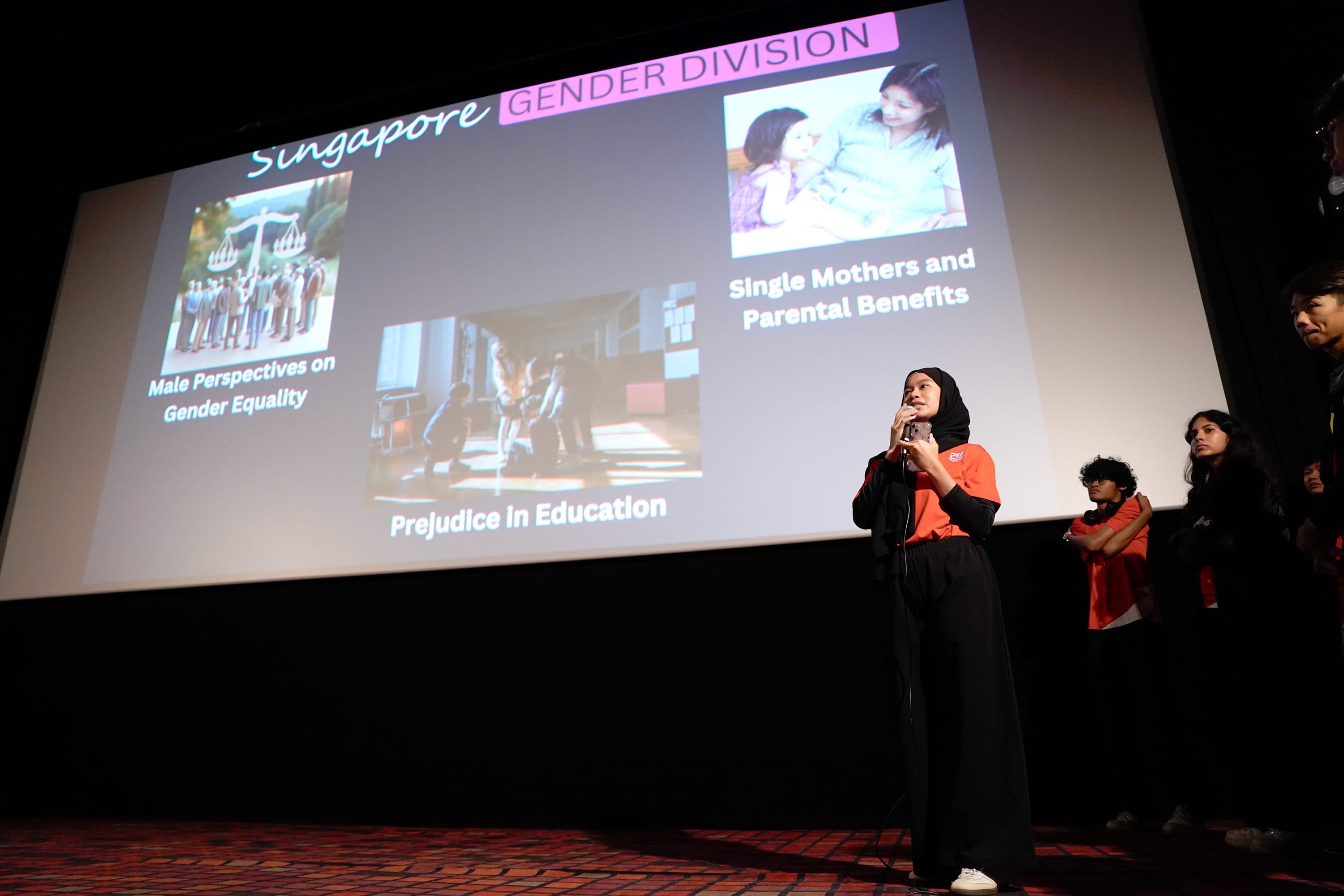A young woman in a hijab speaks into a microphone before a large screen reading "Singapore Gender Division."