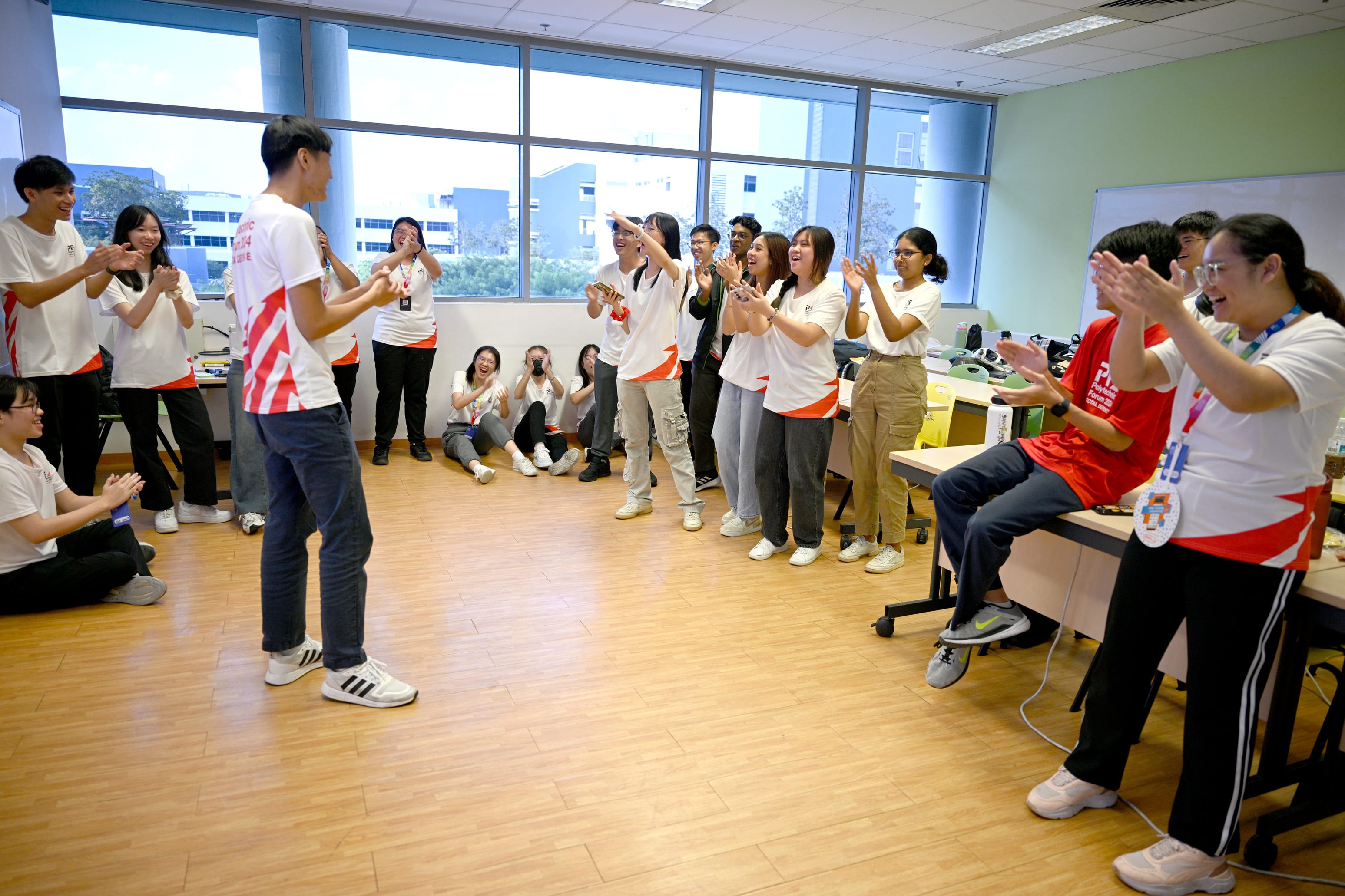 A group of young people in white and red shirts clap and cheer for a student standing in a circle in a classroom.