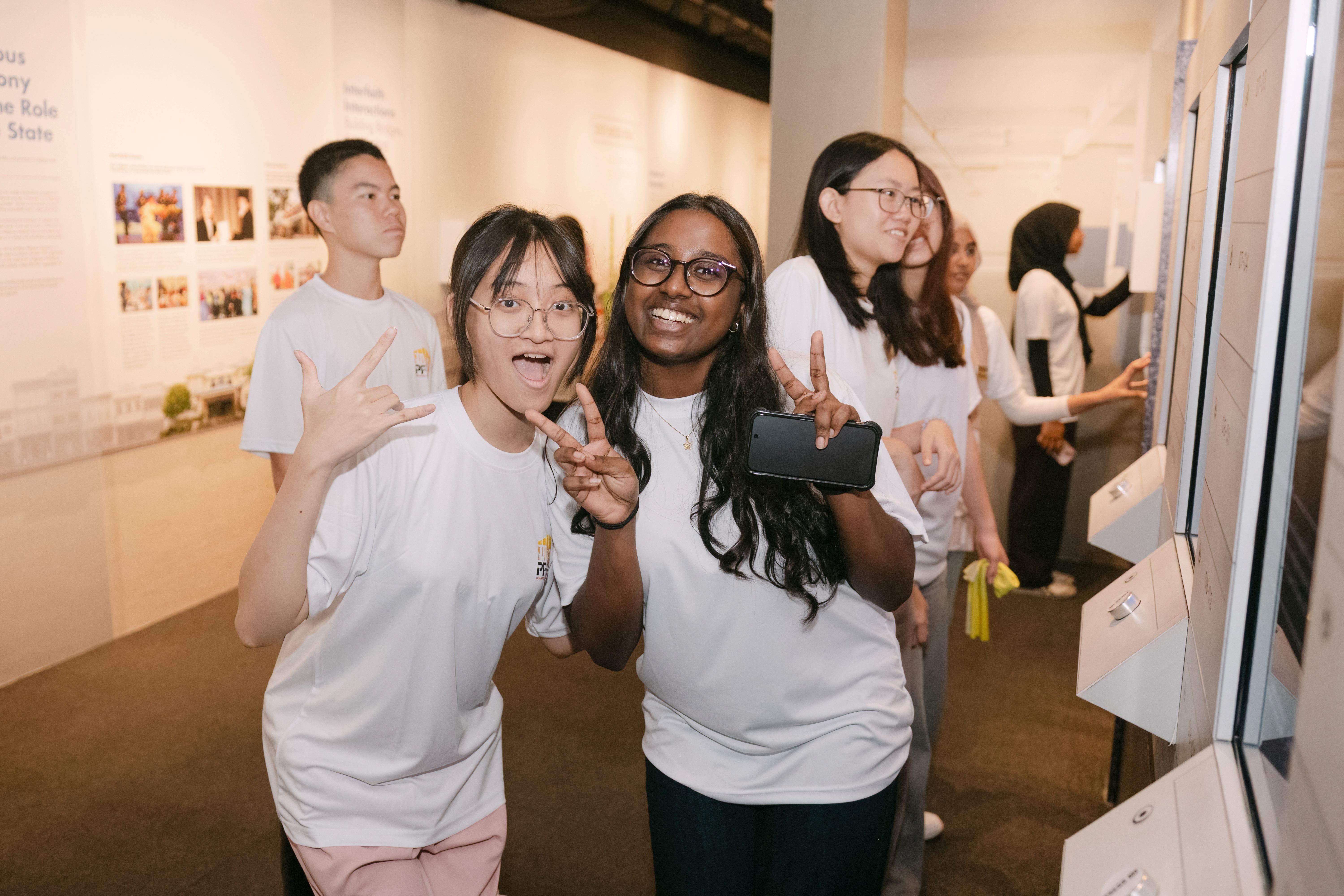 Two smiling young women in white t-shirts pose with peace signs inside a museum exhibition.