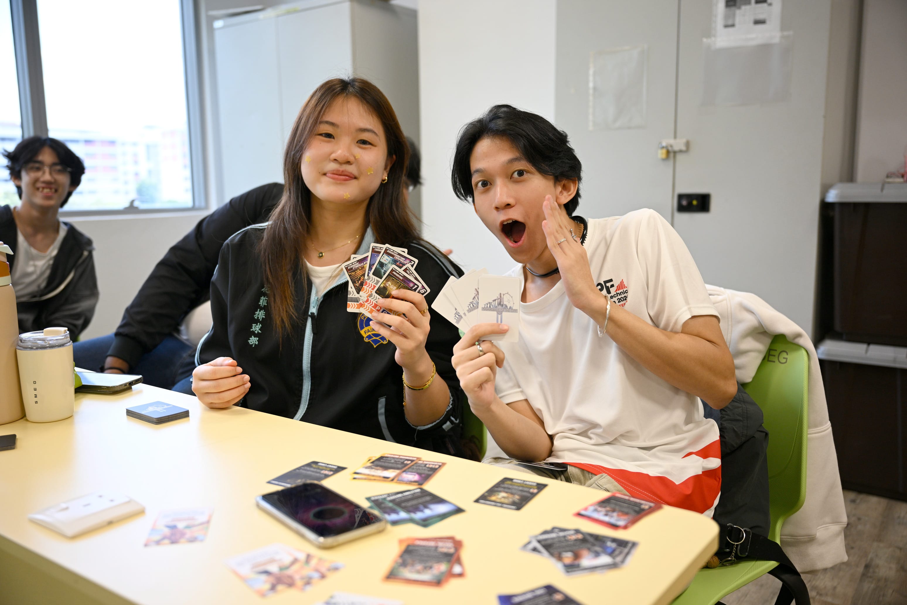 Two young people sit at a table holding trading cards, one smiling and one with a surprised open mouth expression.