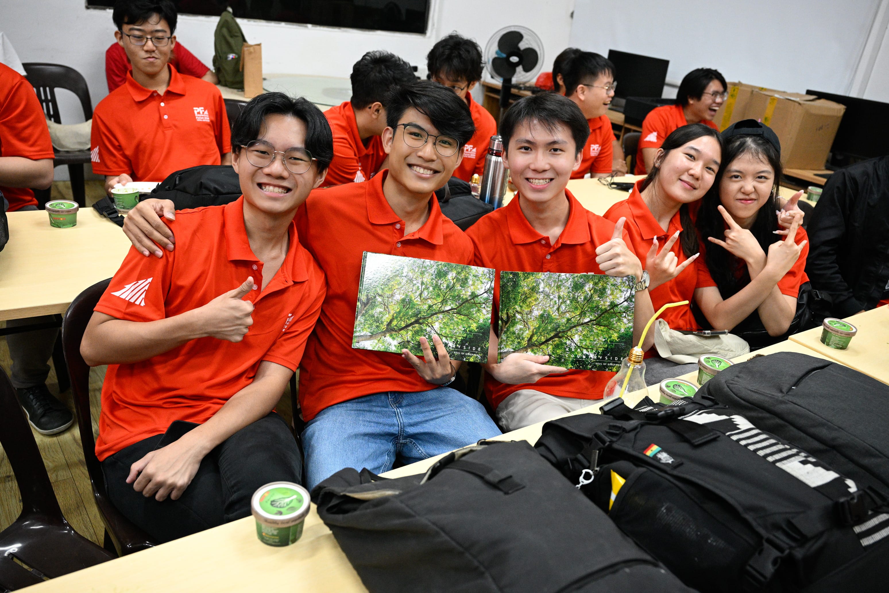 Five smiling young people in matching red polo shirts pose together, two holding photo books showing green trees.