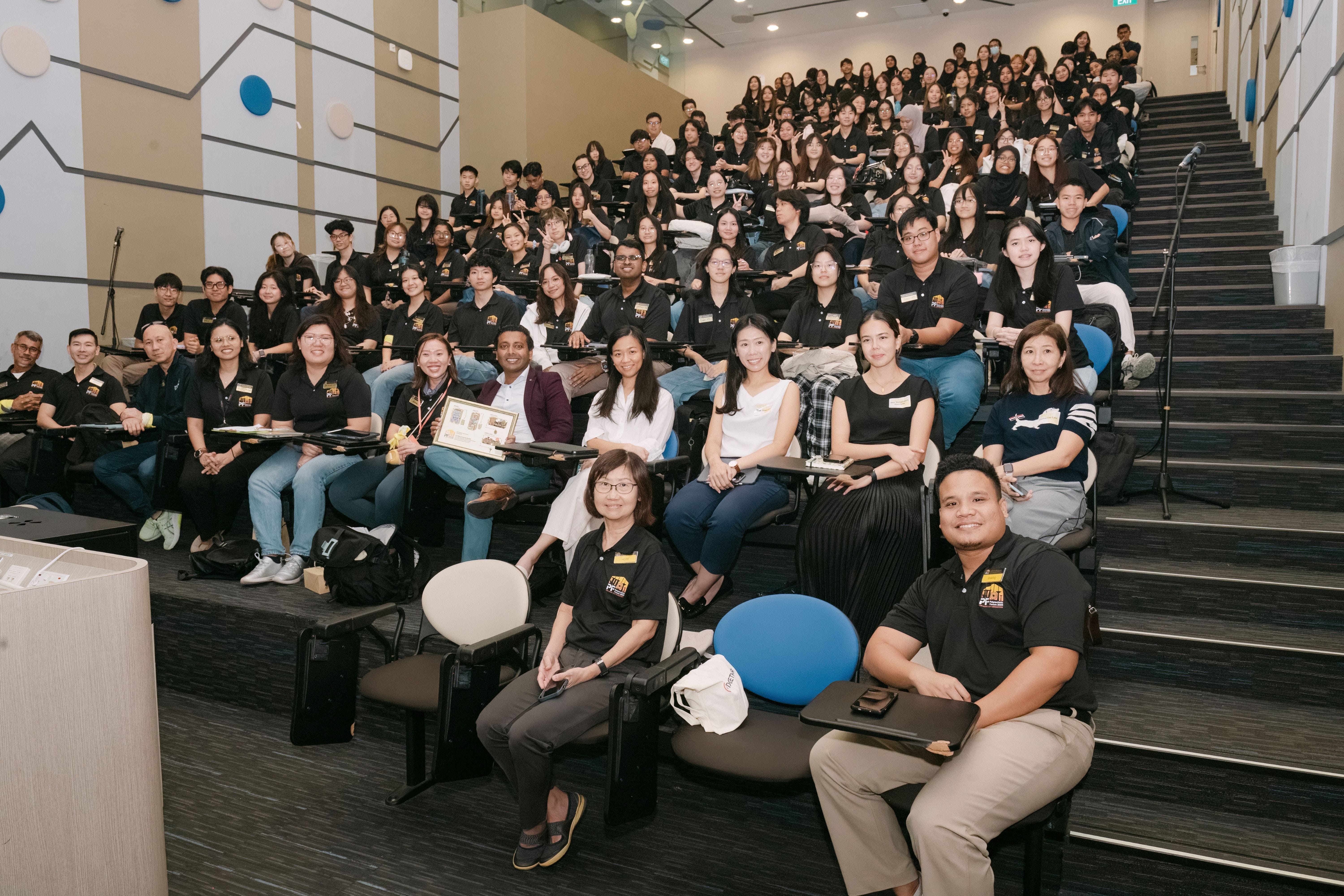 Large group of people mostly wearing black polo shirts seated in a tiered lecture hall, smiling at the camera.