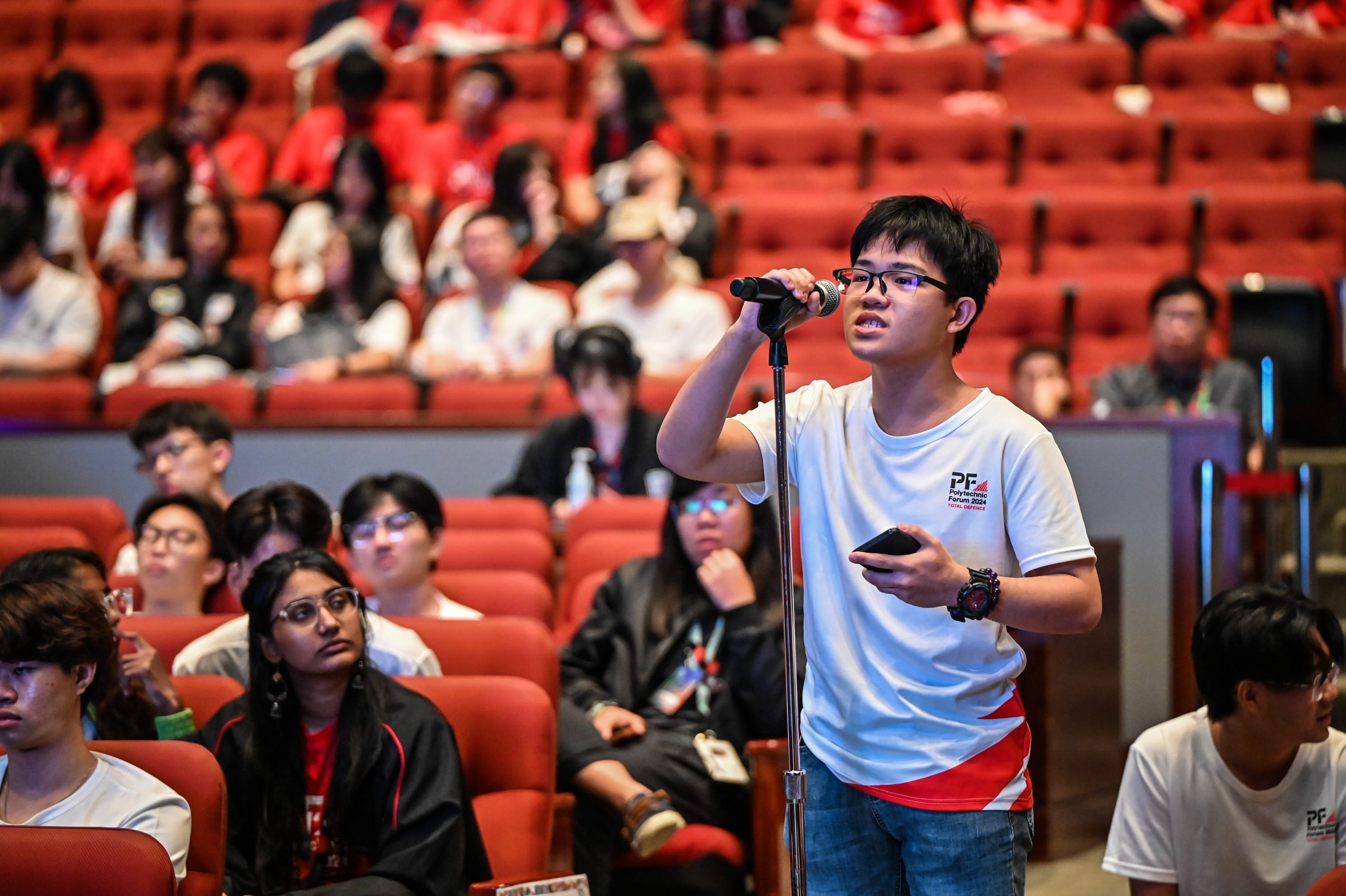 A young man with glasses speaks into a microphone in a red-seated auditorium filled with seated students.