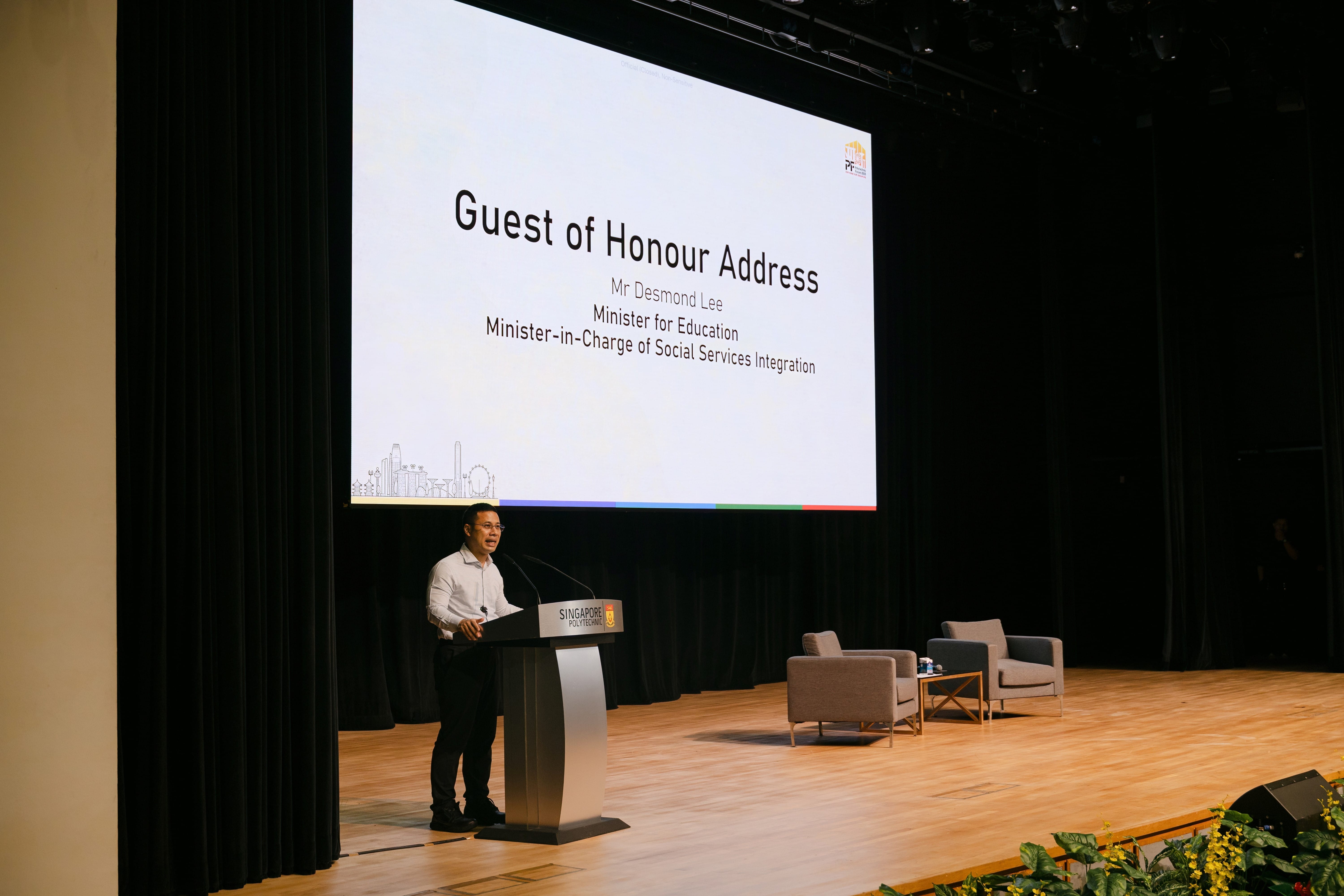 Man speaking at a Singapore Polytechnic podium on stage, with a screen showing "Guest of Honour Address, Mr Desmond Lee."