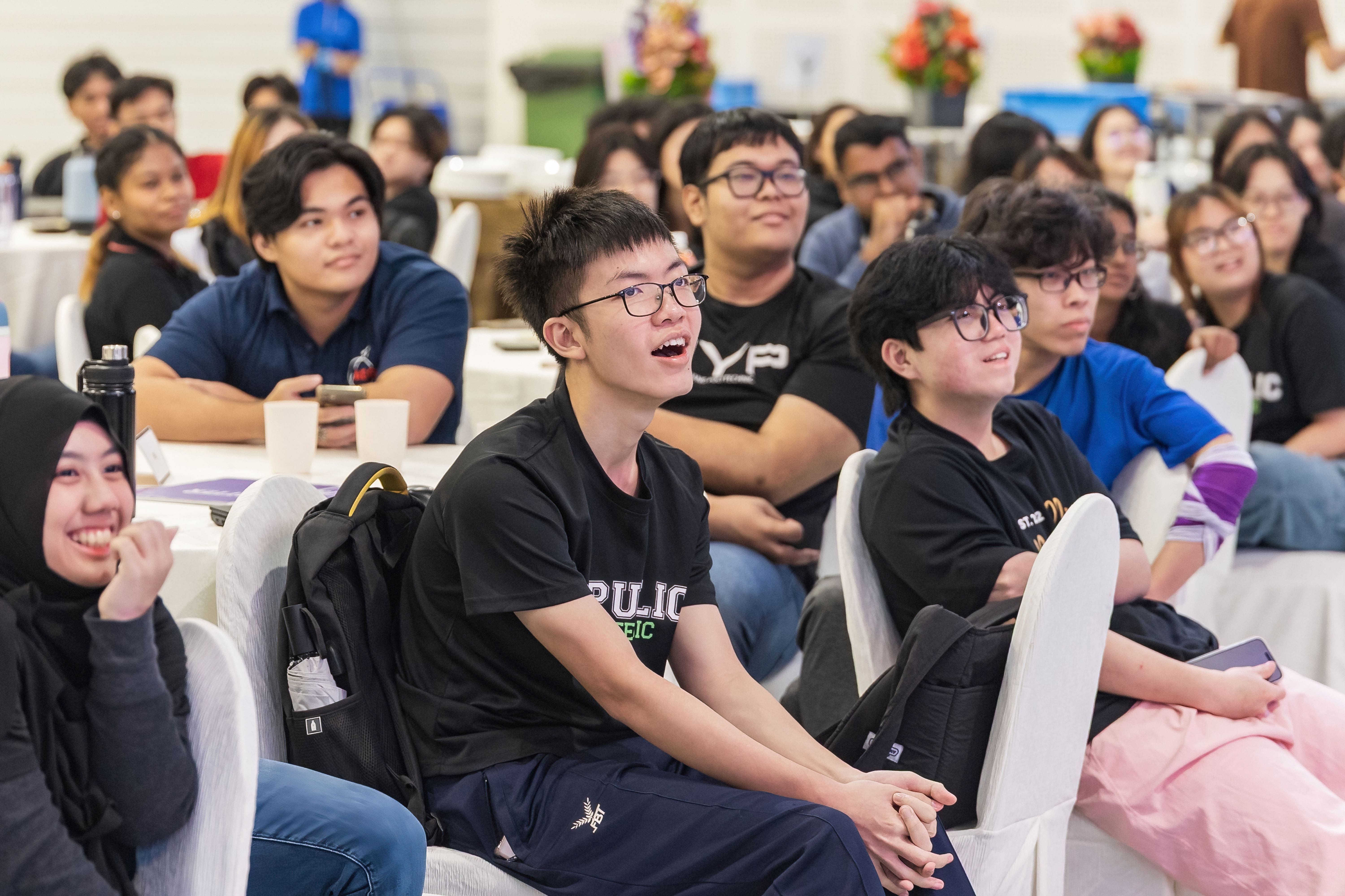 A large group of young people sitting in chairs at an event, smiling and laughing, with floral decorations in the background.
