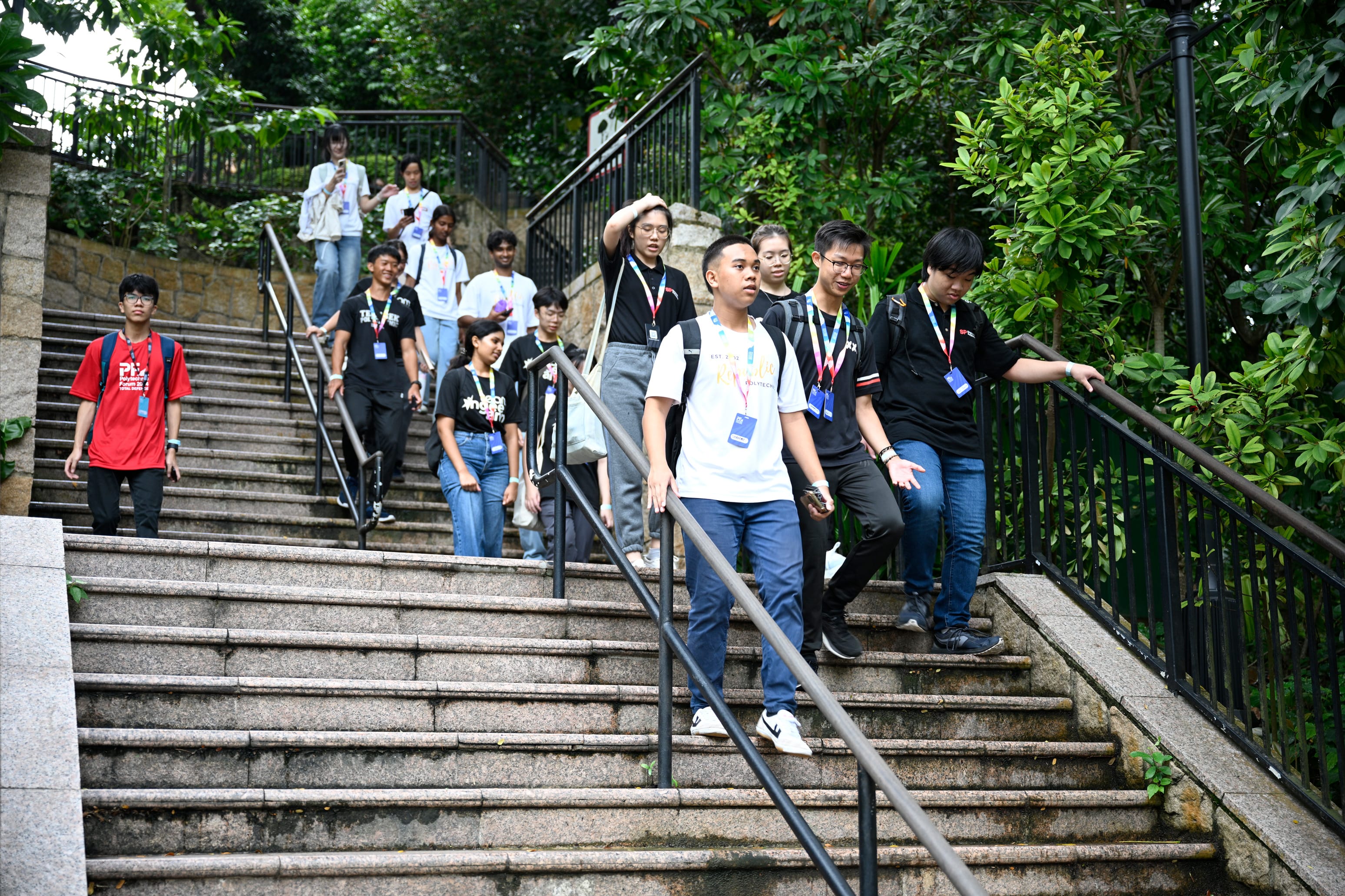 A group of young people wearing lanyards walk down outdoor stone stairs lined with black railings and green trees.