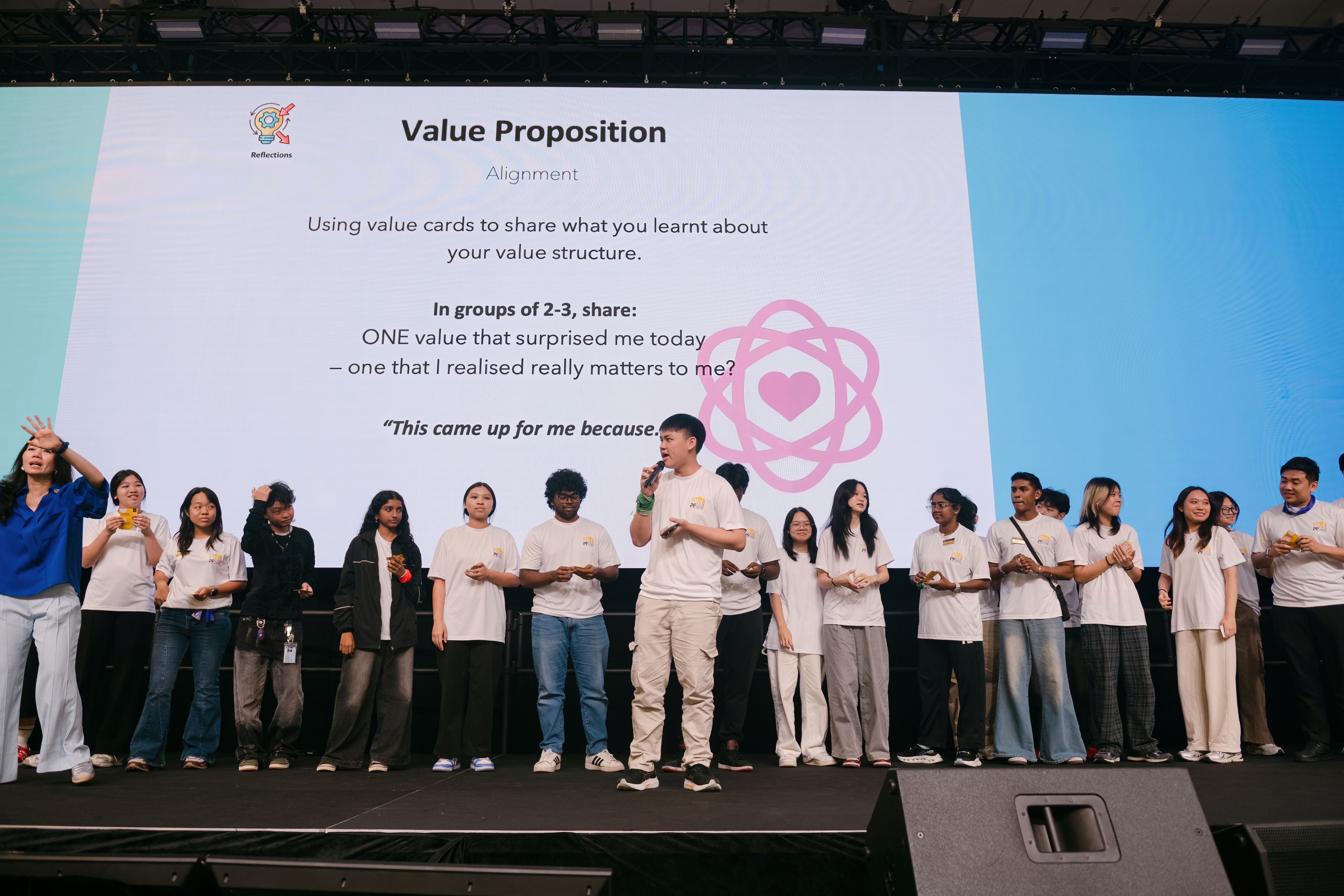 A young man speaks into a microphone on stage with a group in white shirts before a Value Proposition slideshow.