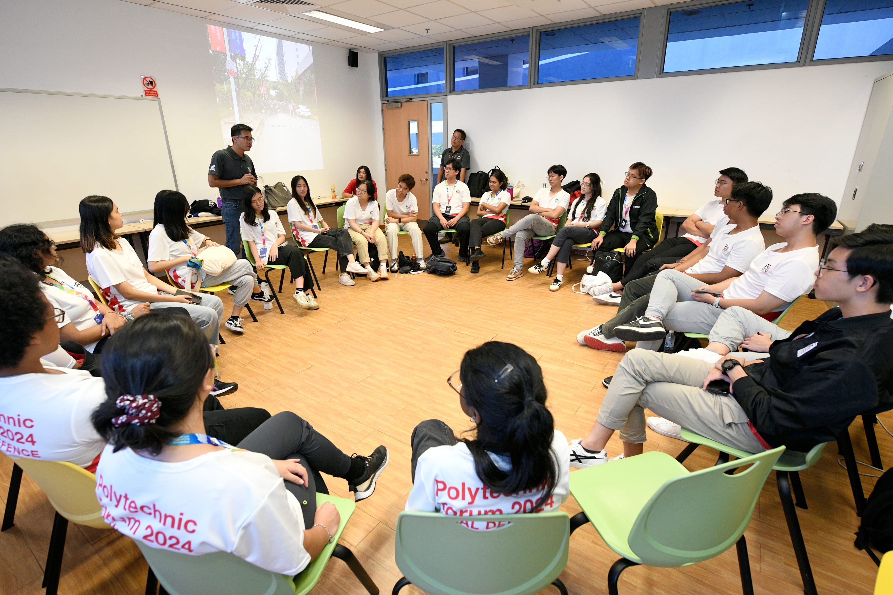 Students in Polytechnic Forum 2024 shirts sit in a circle in a classroom while a man stands and speaks.