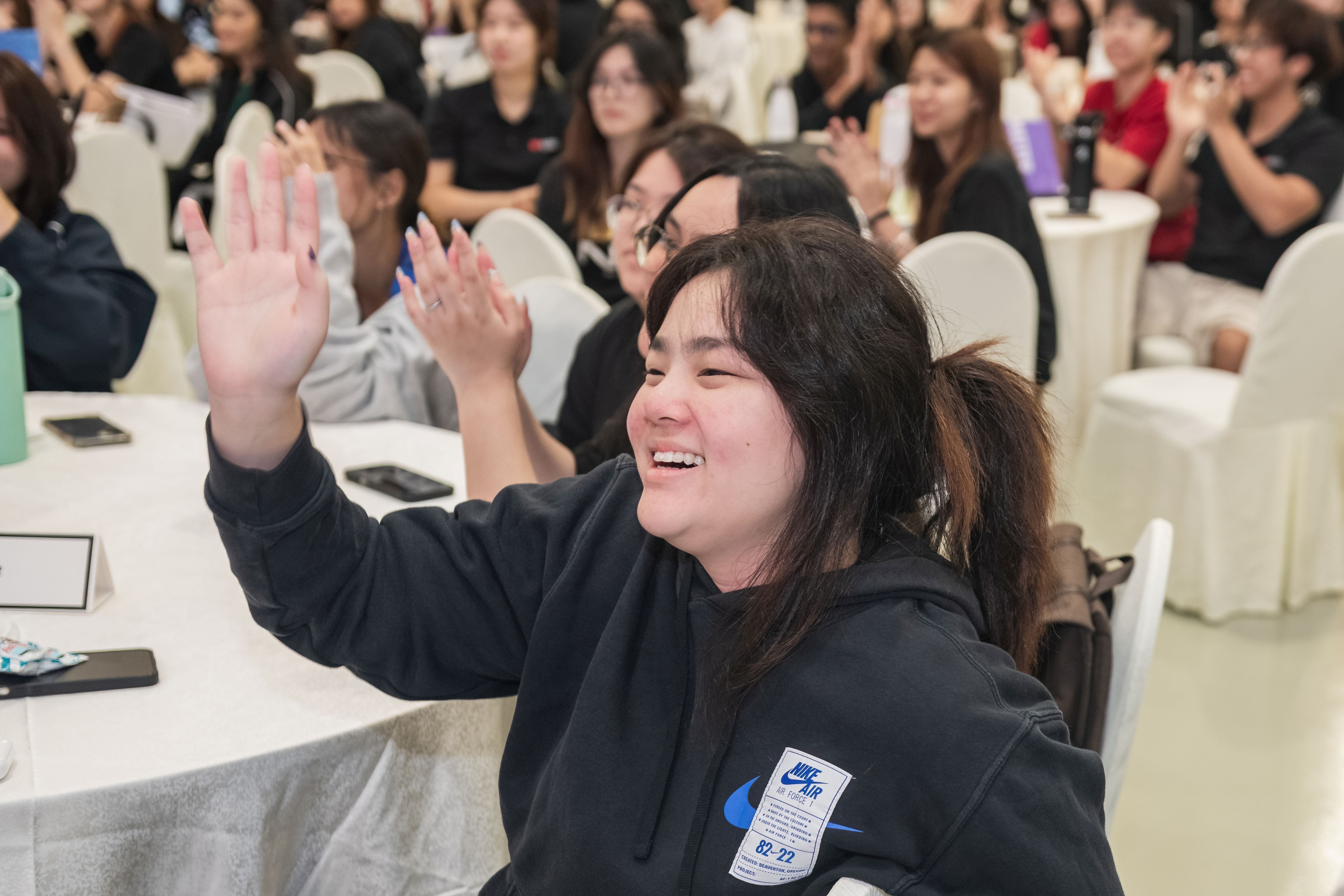 A smiling woman in a black Nike hoodie raises her hand at a crowded seated event with many attendees behind her.